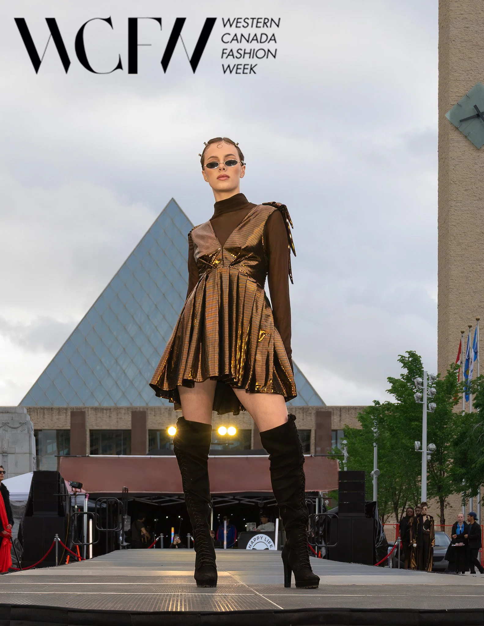 A female model on a runway at Western Canada Fashion Week, wearing a shiny gold and brown dress with black thigh-high boots, sunglasses, and fashion accessories, with an audience and buildings in the background.