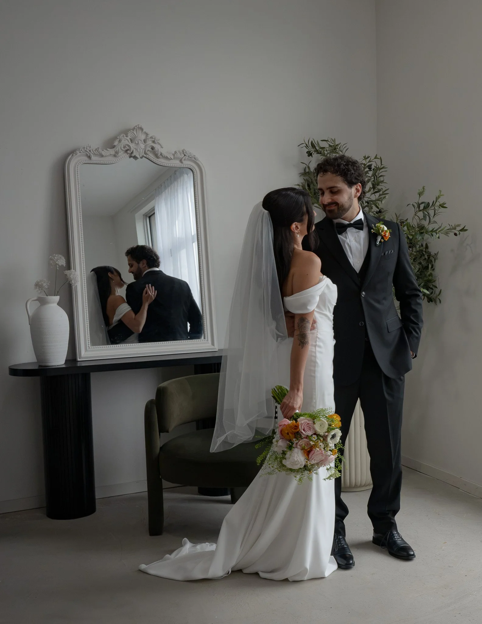 A bride and groom in wedding attire are dancing together indoors, reflected in a large ornate mirror on the wall behind them. The bride wears a white dress, and the groom wears a black tuxedo with a bow tie. The bride is holding a bouquet of flowers,
