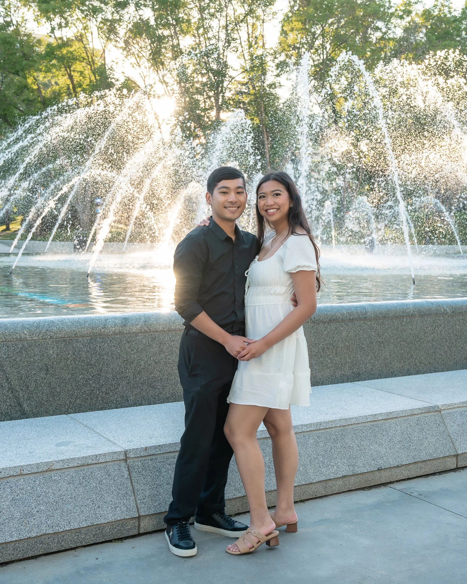 A happy couple standing in front of a water fountain with tall trees in the background during sunset. The man is wearing a black shirt and black pants, while the woman is dressed in a white dress and tan heeled sandals.