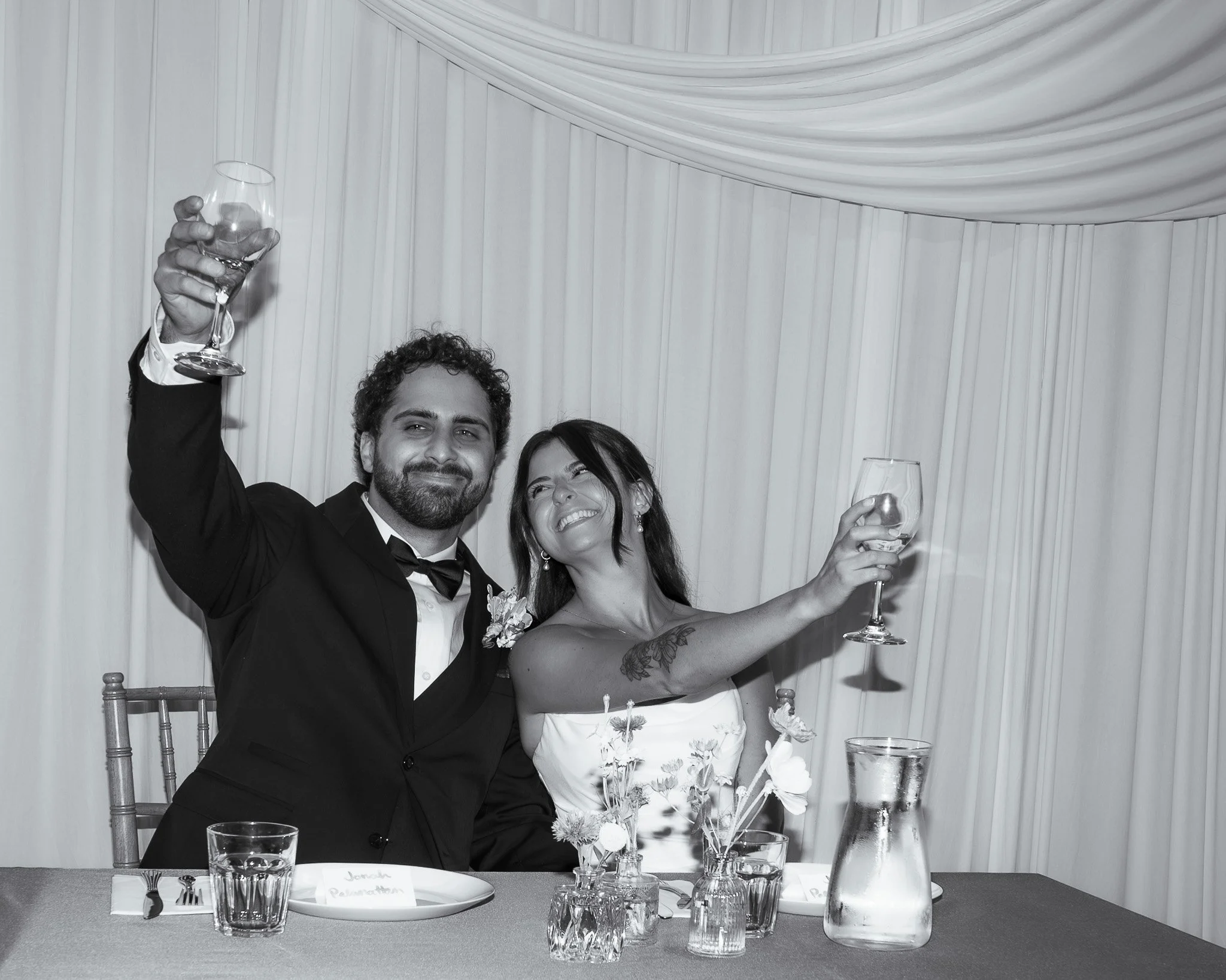Two people celebrating at a formal event, raising glasses of wine. The man is dressed in a tuxedo, and the woman is wearing a strapless dress with a floral pattern. They are smiling, and the setting includes a table with glasses and flower arrangemen
