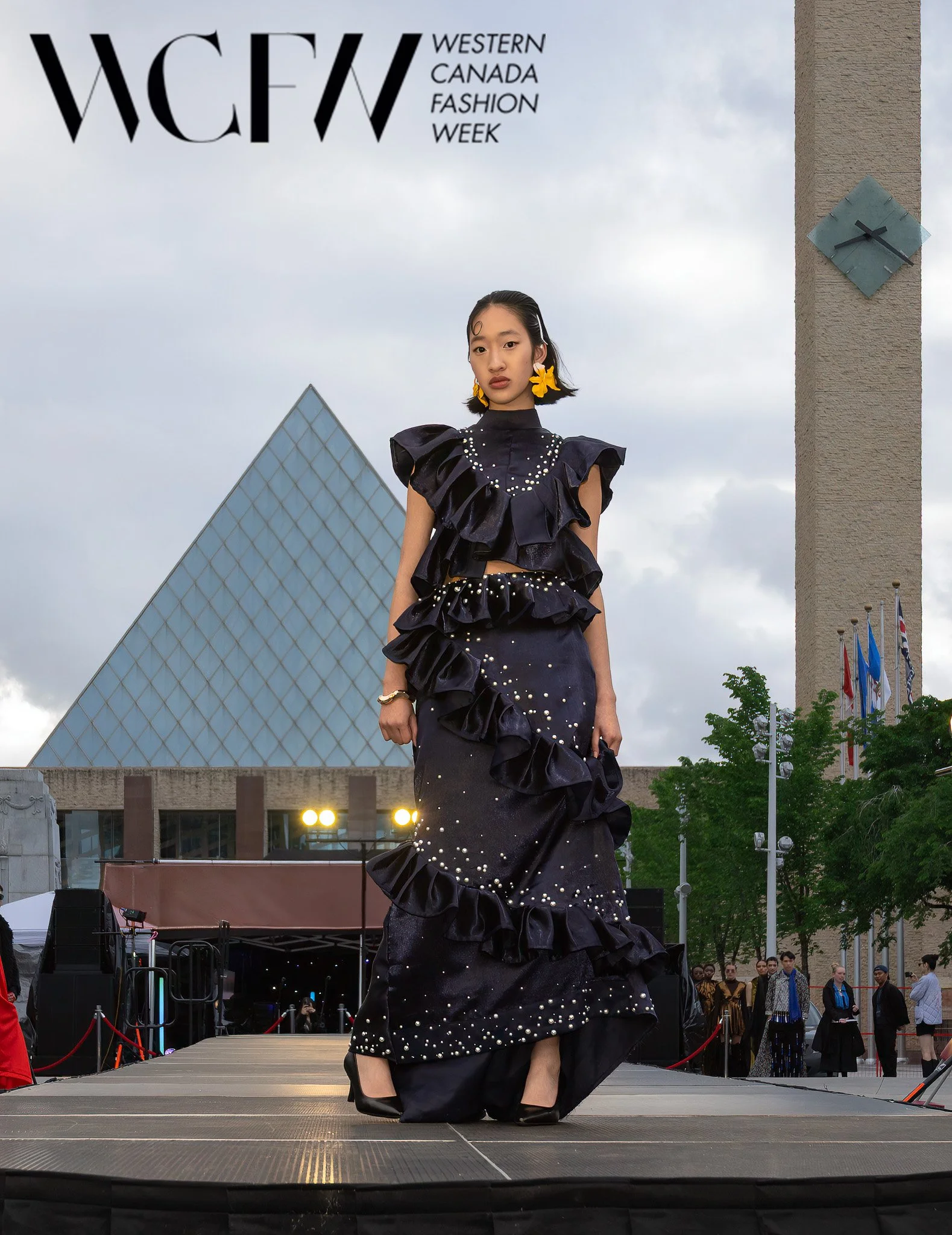 A woman walking on a runway at Western Canada Fashion Week, wearing a dramatic black dress with ruffled layers and pearl embellishments, with a crowd and flags in the background.