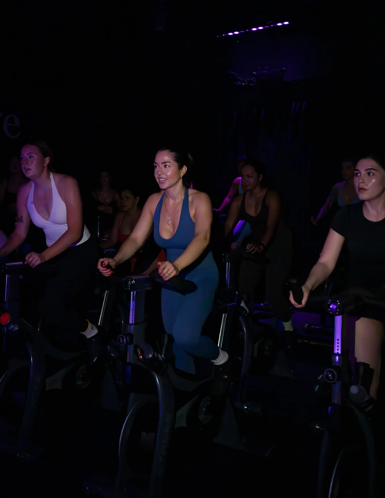 People participating in a dark indoor cycling class, illuminated by purple and blue lighting, riding stationary bikes