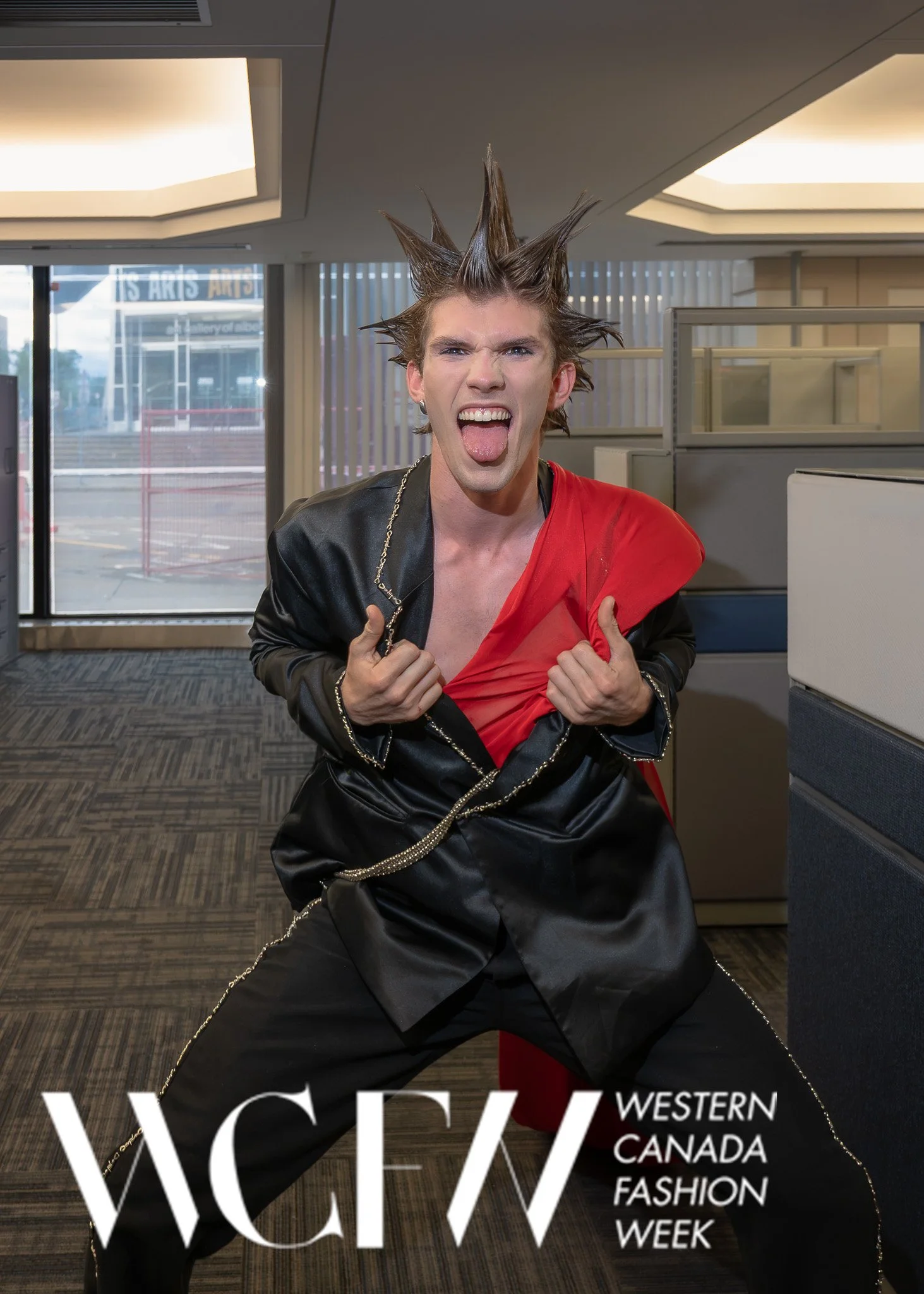 Person with spiked hair sticking out in all directions, wearing a black and red outfit, sticking out tongue, inside modern indoor space with large windows and government building in background, promotional text for Western Canada Fashion Week