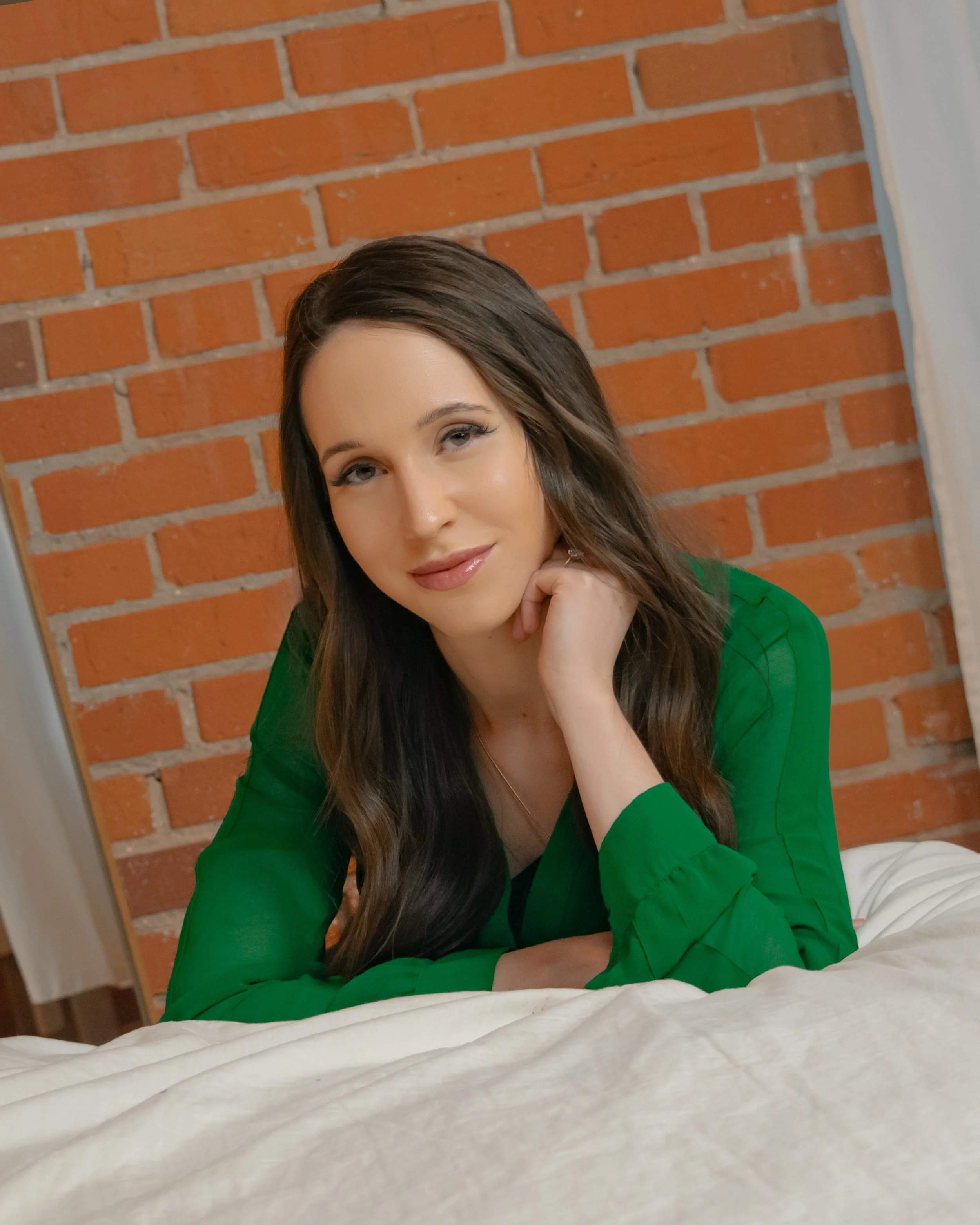 A woman with long brown hair, wearing a green top, lying on a bed with a beige bedspread, resting her chin on her hand, in front of an orange brick wall.
