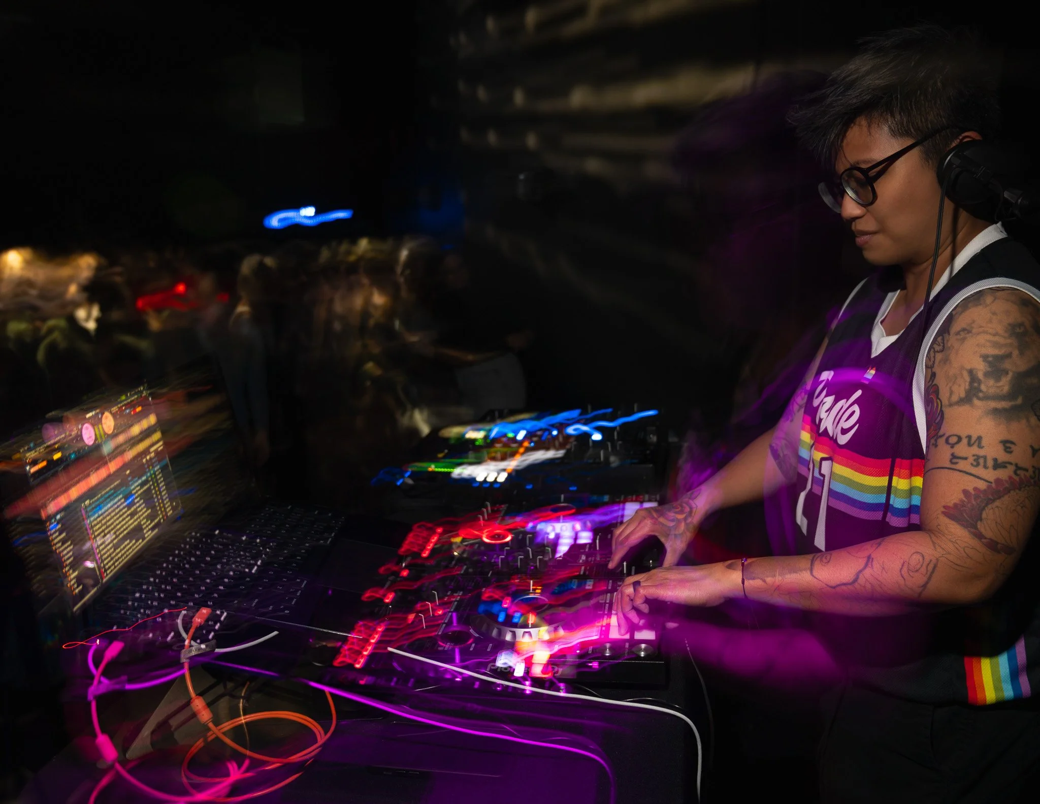 A female DJ with tattoos on her arms, wearing glasses and a purple tie-dye shirt, operating a DJ mixer with colorful lights in a dark setting.