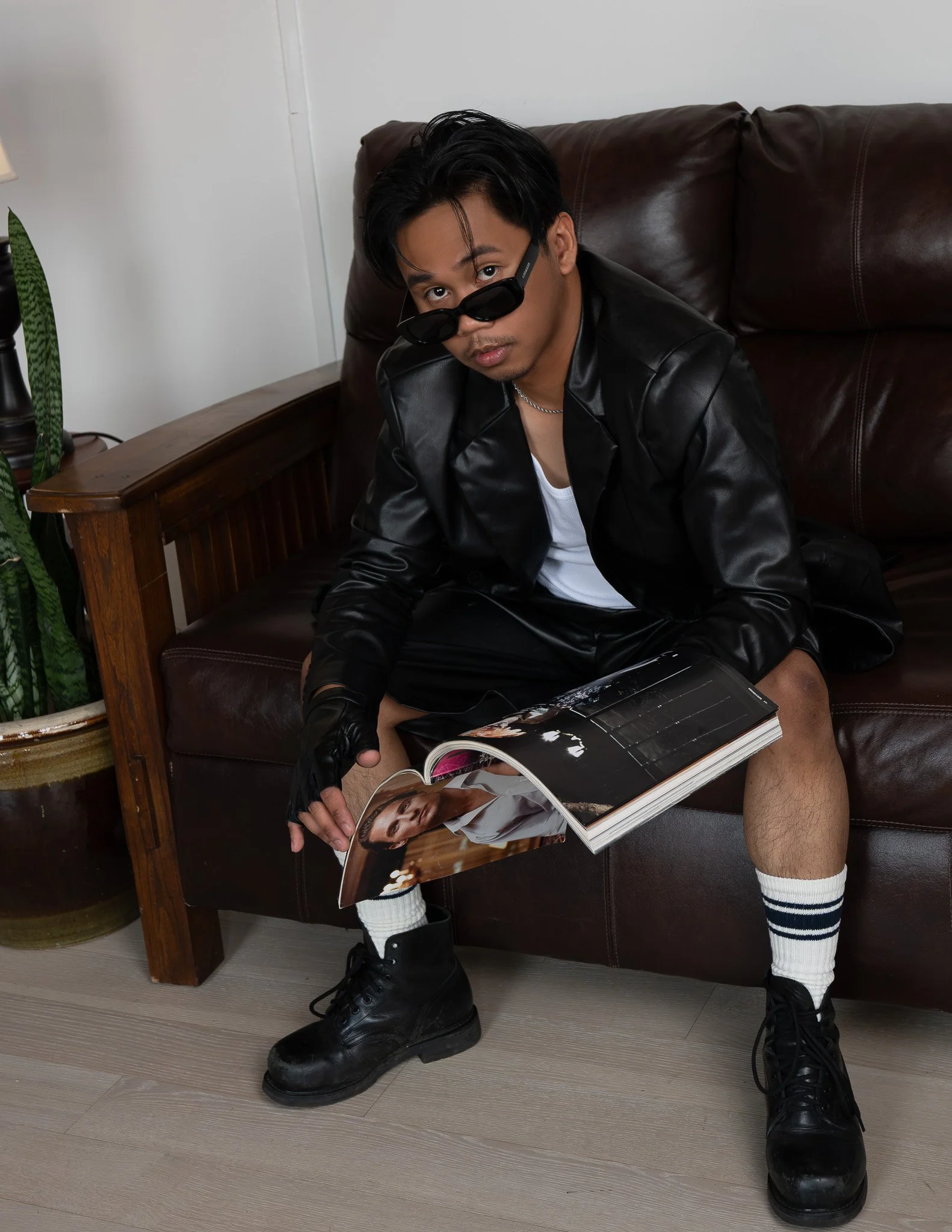 A young man sitting on a brown leather couch, wearing black leather jacket, white shirt, black boots, white knee-high socks with black stripes, sunglasses, and looking towards the camera while holding a magazine.