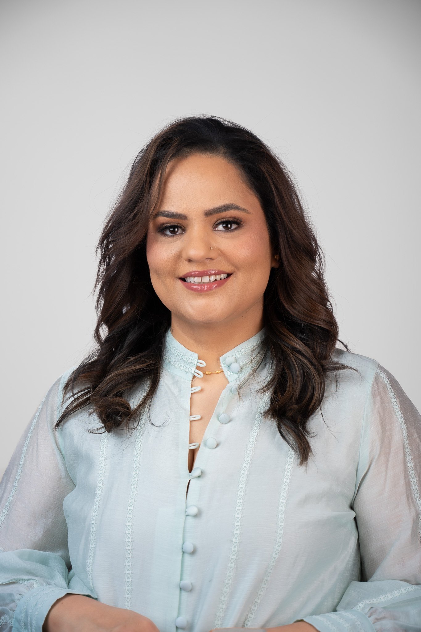 A woman with dark wavy hair and makeup, wearing a light blue blouse with lace details and buttons, smiling against a plain gray background.