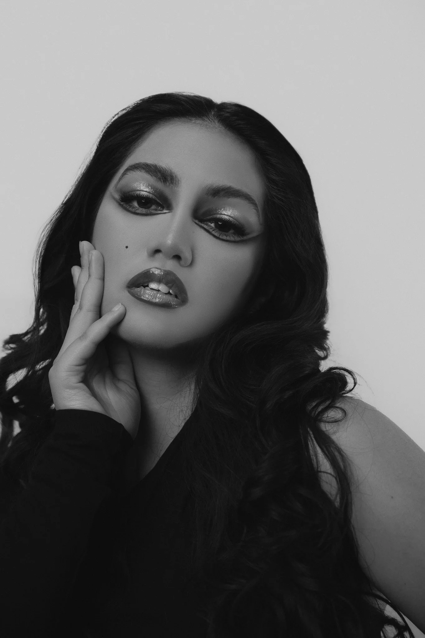 A black and white portrait of a woman with long, wavy hair, bold makeup, and a contemplative pose, with her hand touching her face.