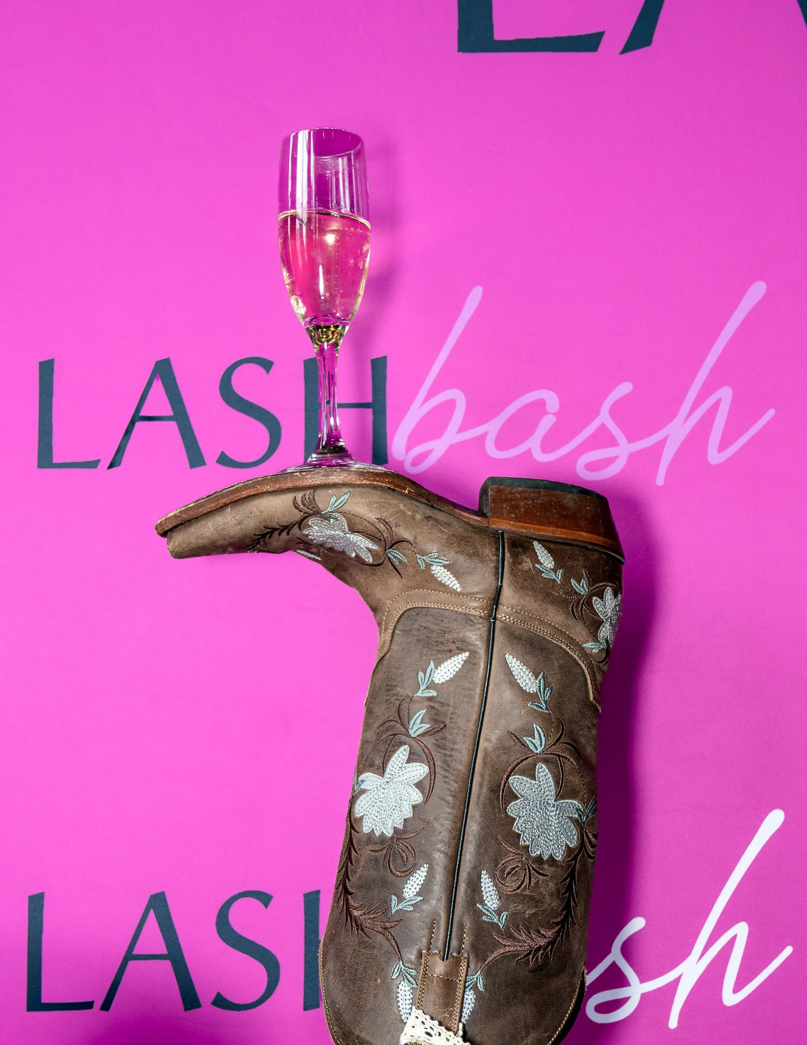 A high-heeled cowboy boot with floral embroidery, placed on a pink surface with the words 'LASH' and 'lash' in the background.