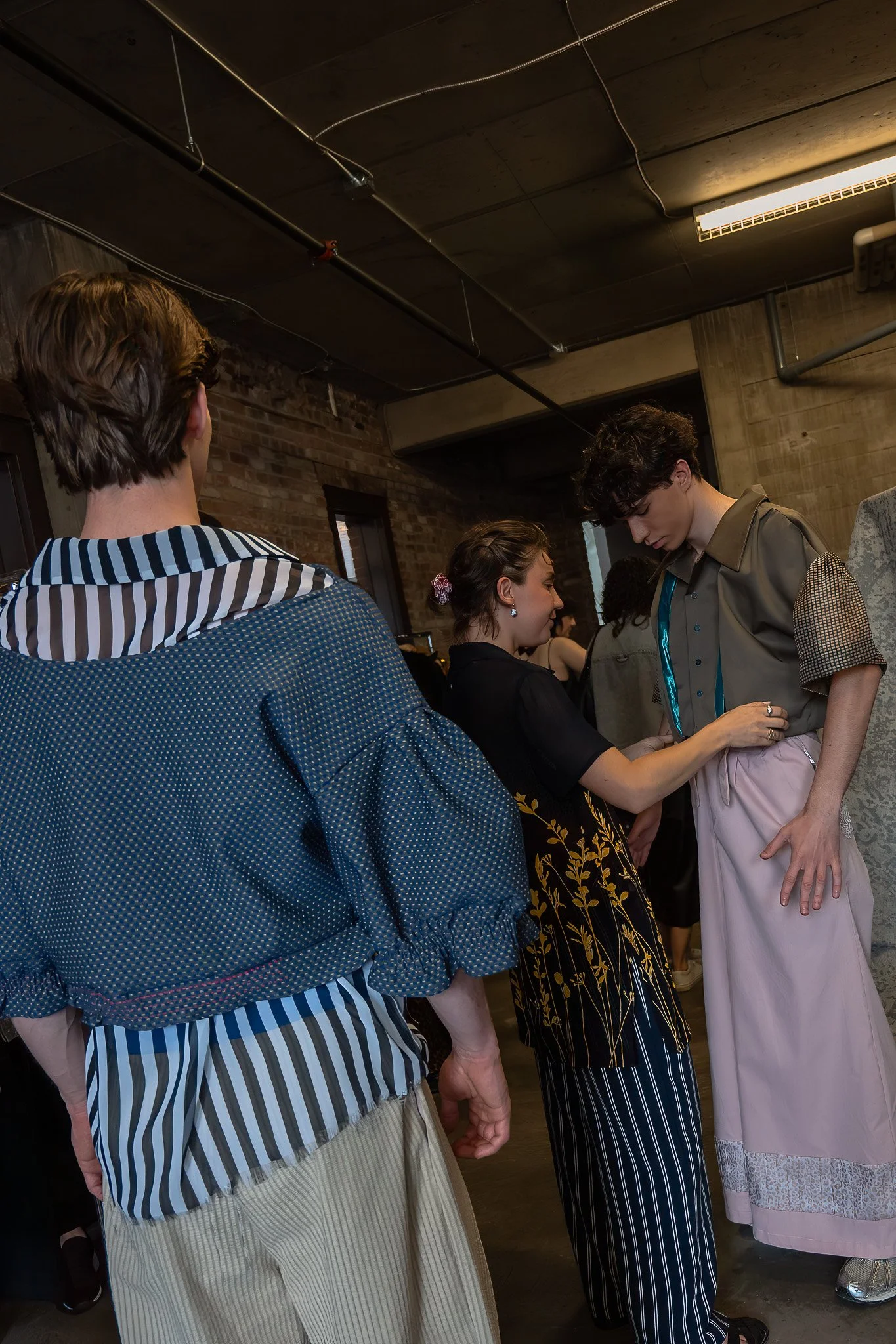 People standing close together, with two women facing each other, talking. The scene is indoors with a brick wall and industrial ceiling lights.