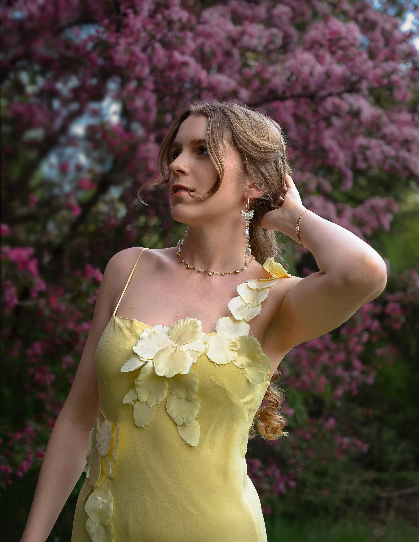 A woman with long, light brown hair, wearing a yellow dress with decorative flowers, standing in front of a pink flowering tree. She is posing with one hand behind her head and looking to the side, adorned with earrings and a necklace.