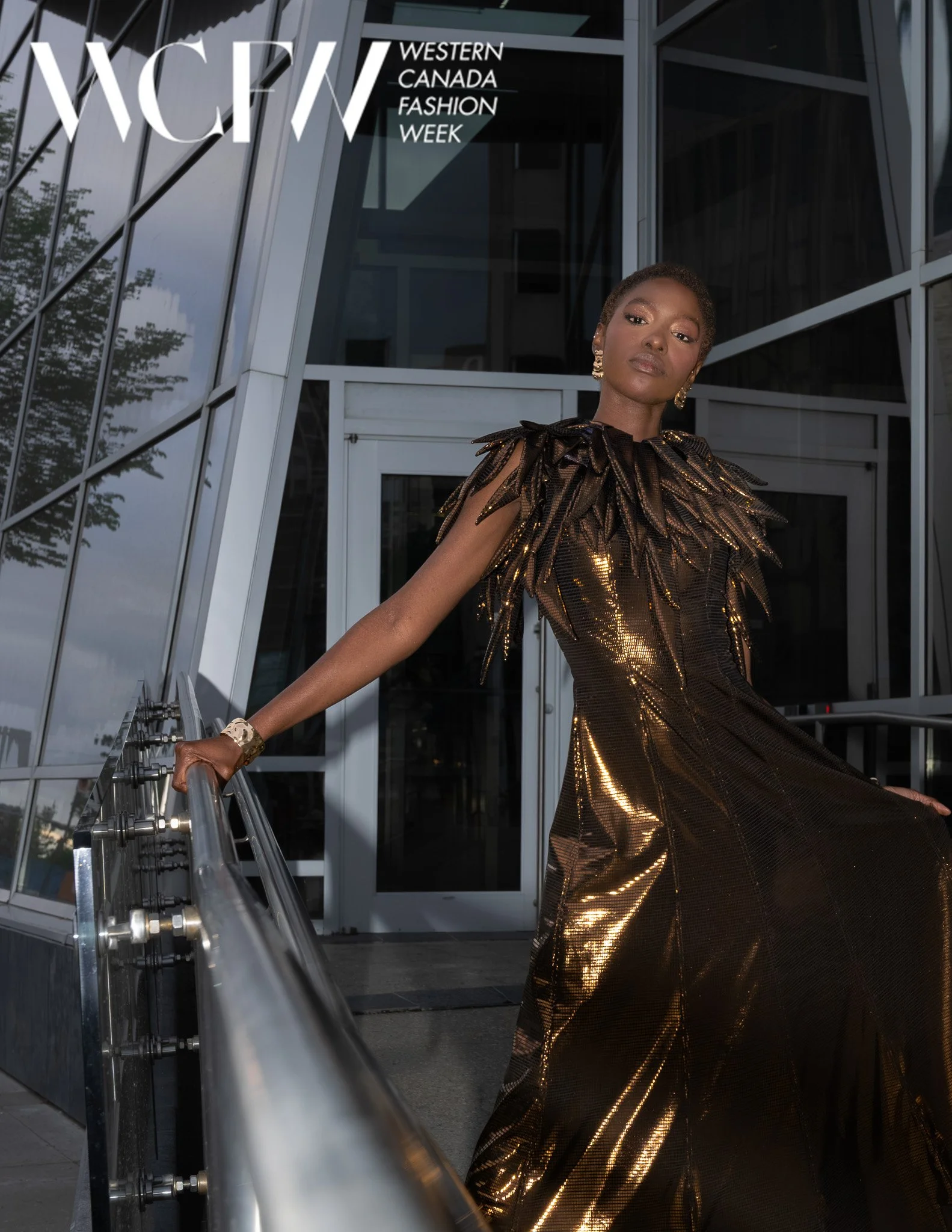 A woman in a gold, metallic dress with feather-like details stands on a modern glass and metal staircase outside a building with architecturally styled glass walls. The scene is part of Western Canada Fashion Week.