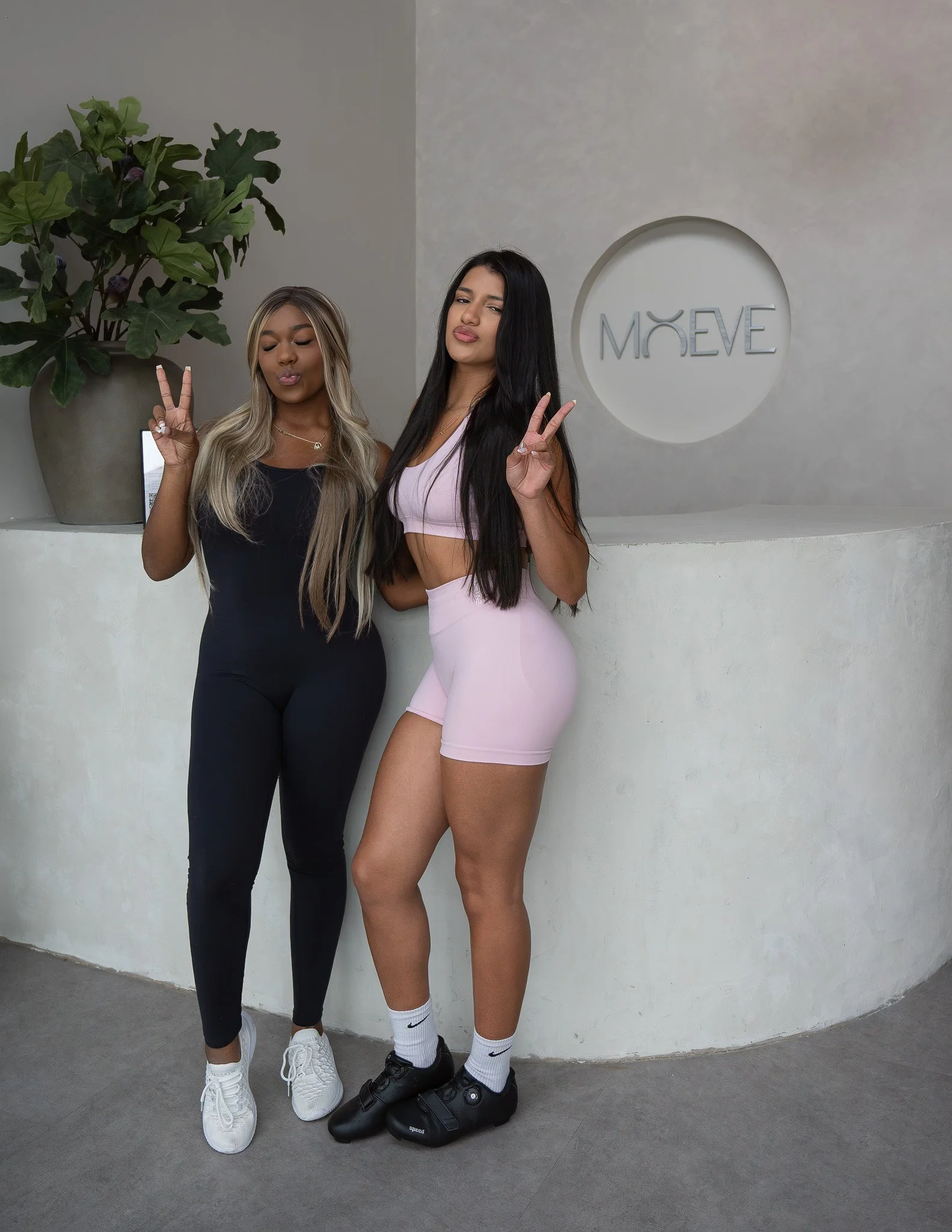 Two women standing in front of a white wall with a round sign that reads "MÖVE". The woman on the left has long blonde hair, dark skin, and is wearing a black sports outfit, white sneakers, and is making a peace sign with her right hand. The woman on