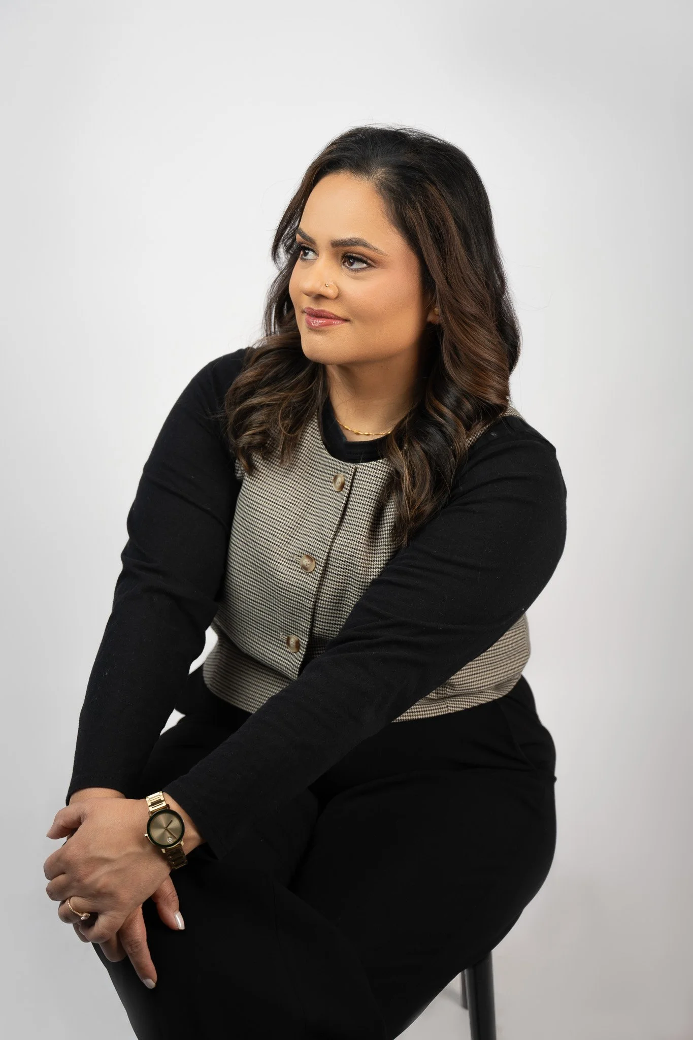 A woman with dark brown wavy hair, wearing a black and beige checkered vest over a black long-sleeve top, is sitting on a black chair against a plain white background. She has a gold watch, a small nose piercing, and a delicate gold necklace. She is 