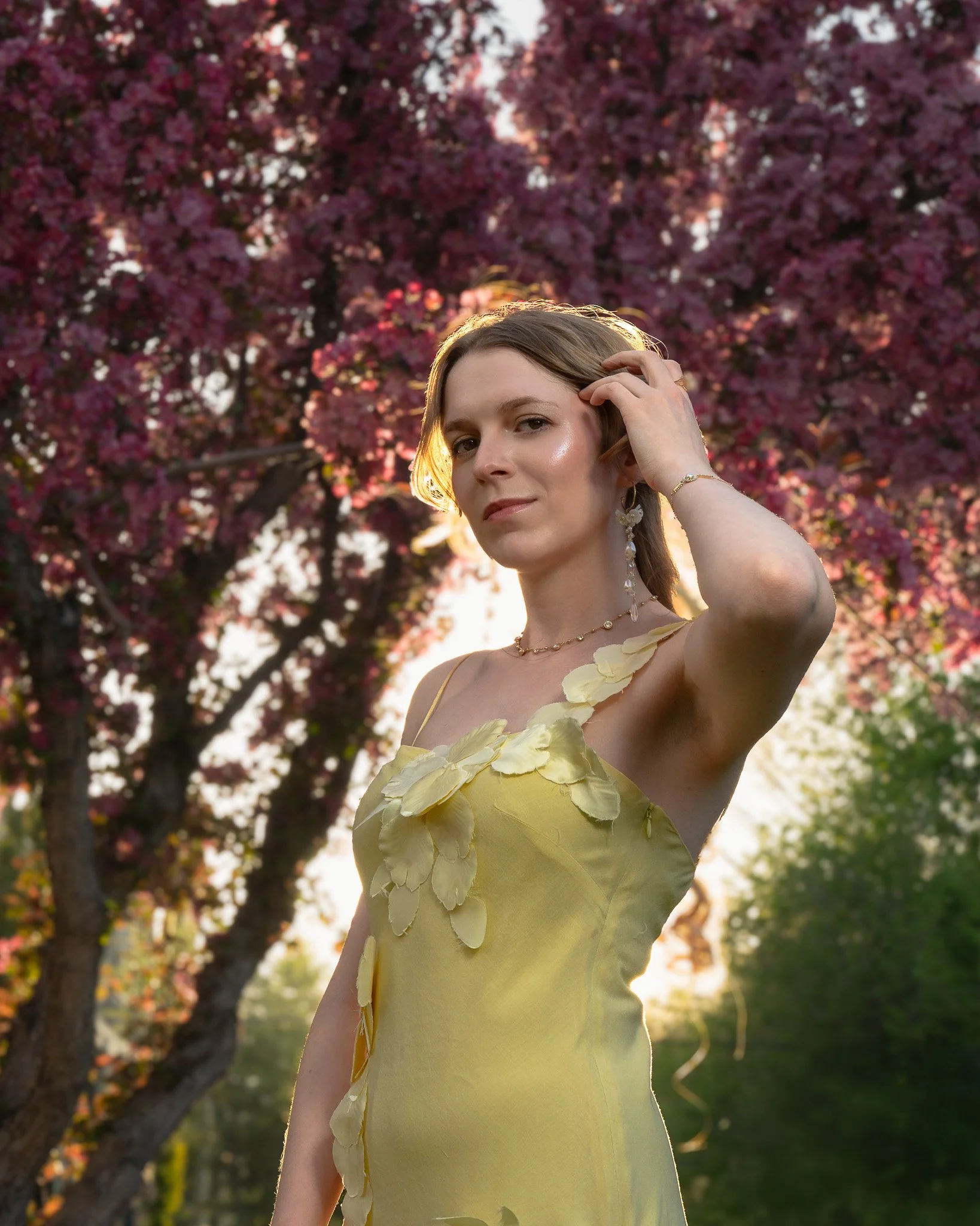 A woman in a yellow dress with floral embellishments, standing outdoors at sunset with pink flowering trees in the background.