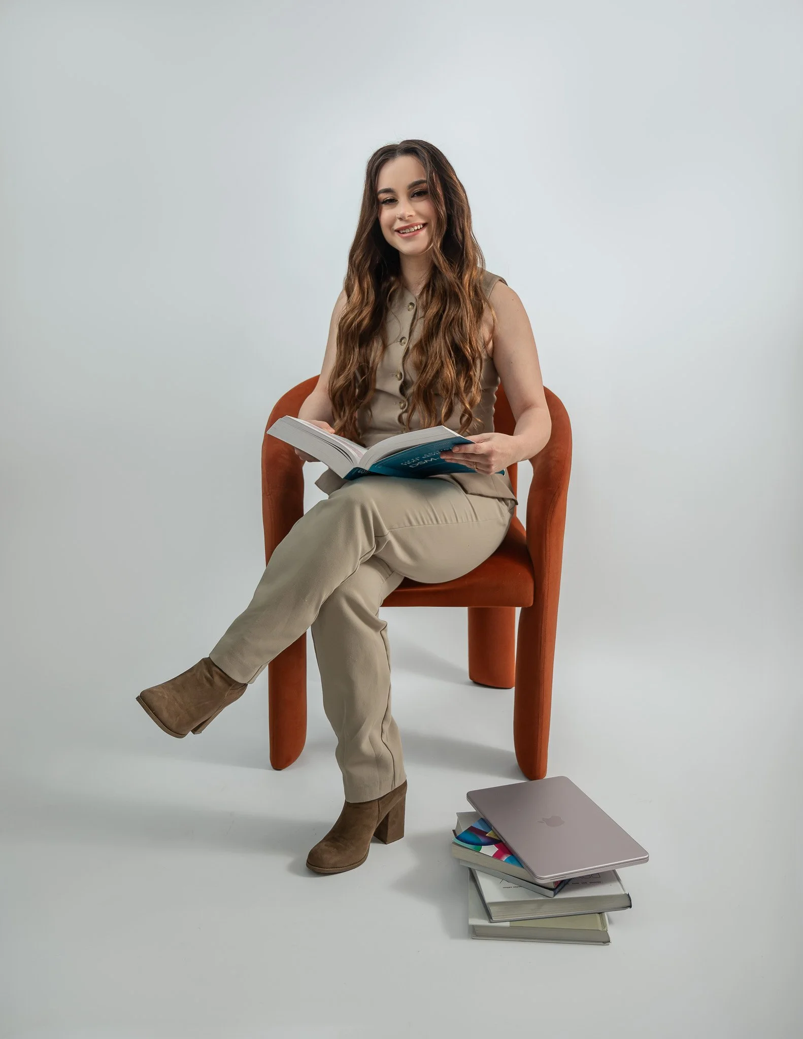 Young woman with long wavy hair, smiling, sitting in an orange chair, reading a book, wearing a beige sleeveless top and beige pants, with a laptop and stacked books on the floor nearby, against a plain light gray background.
