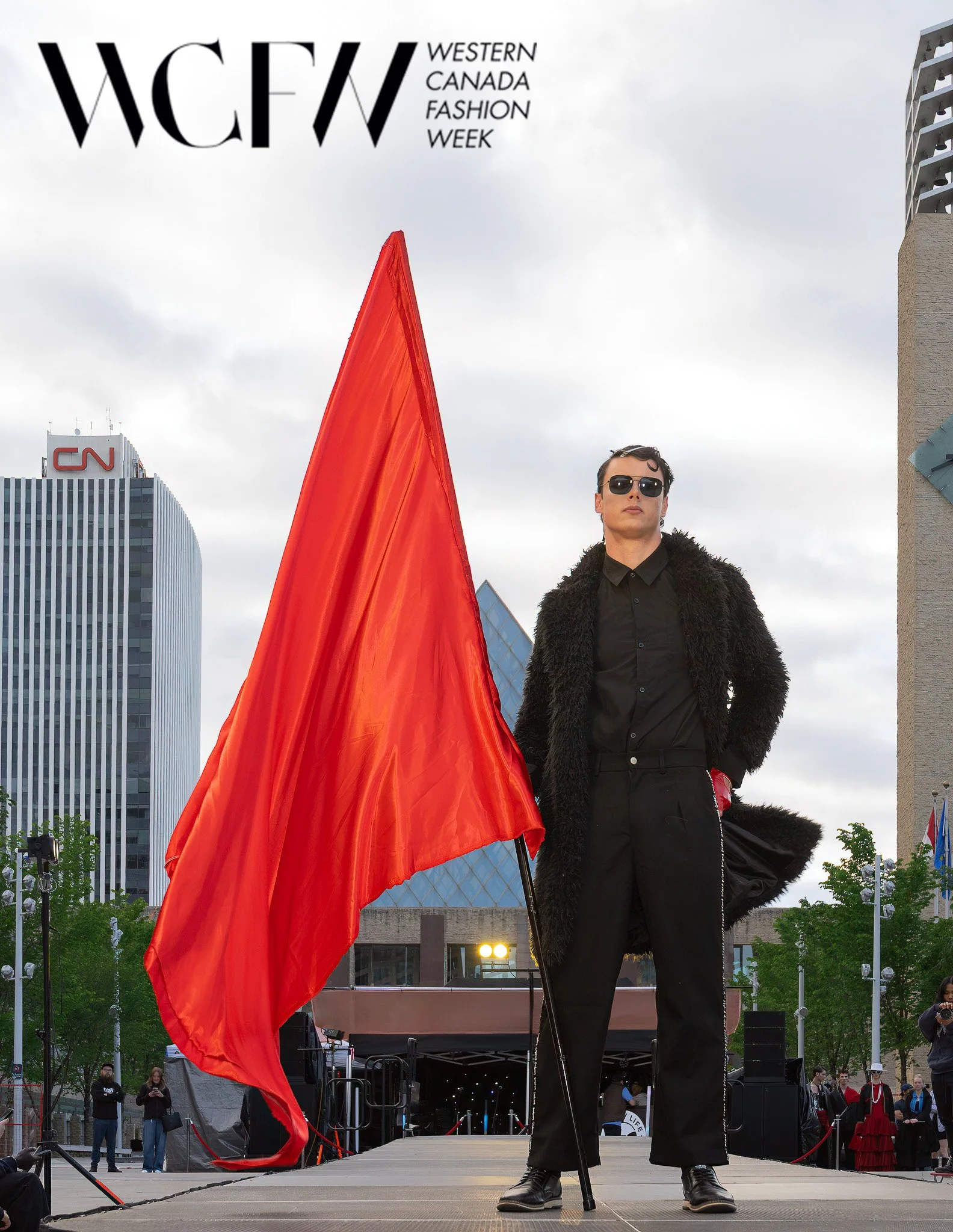 A person dressed in black with sunglasses and a furry coat stands on a stage at Western Canada Fashion Week, holding a large red flag in front of a cityscape with tall buildings, including a CNN building, and a cloudy sky.