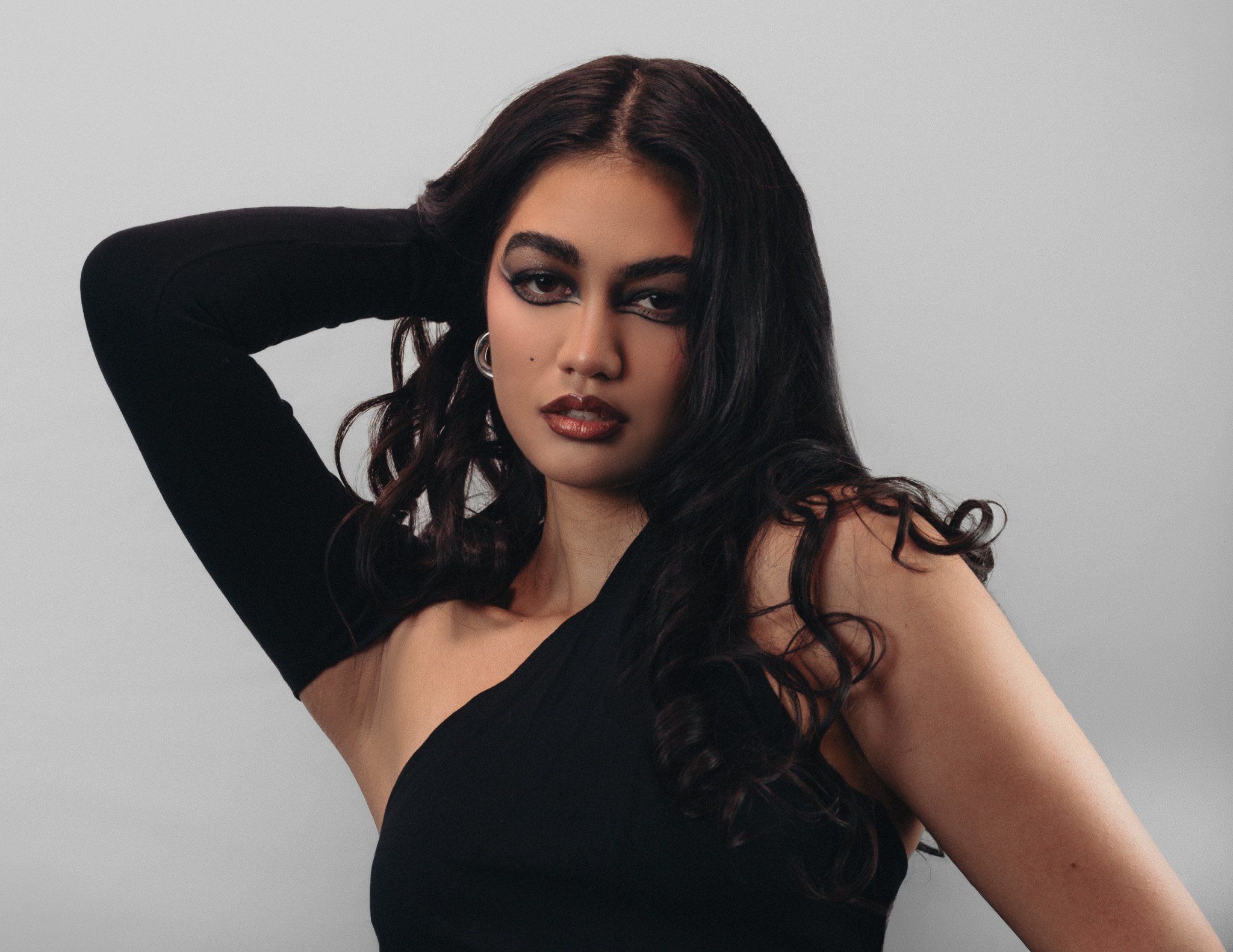 A woman with dark wavy hair wearing a black top and heavy makeup, posing with one arm bent behind her head against a plain background.