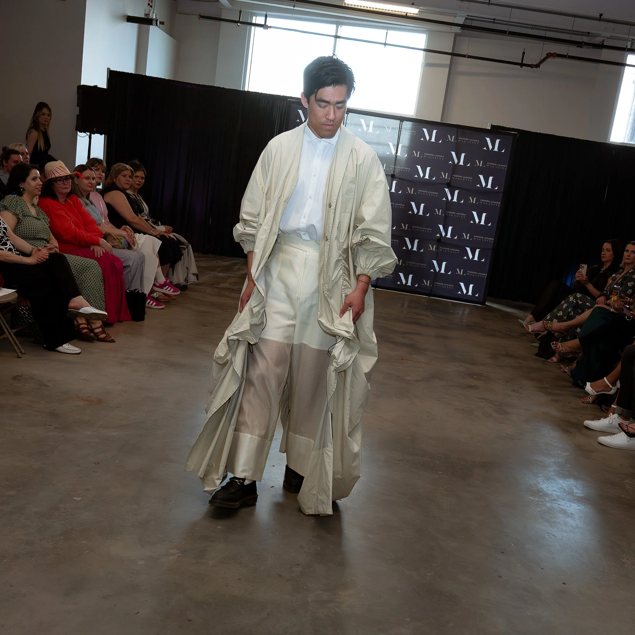 A male model walking on a runway during a fashion show, wearing a cream jacket and wide cream pants, with an audience seated on either side of the runway.