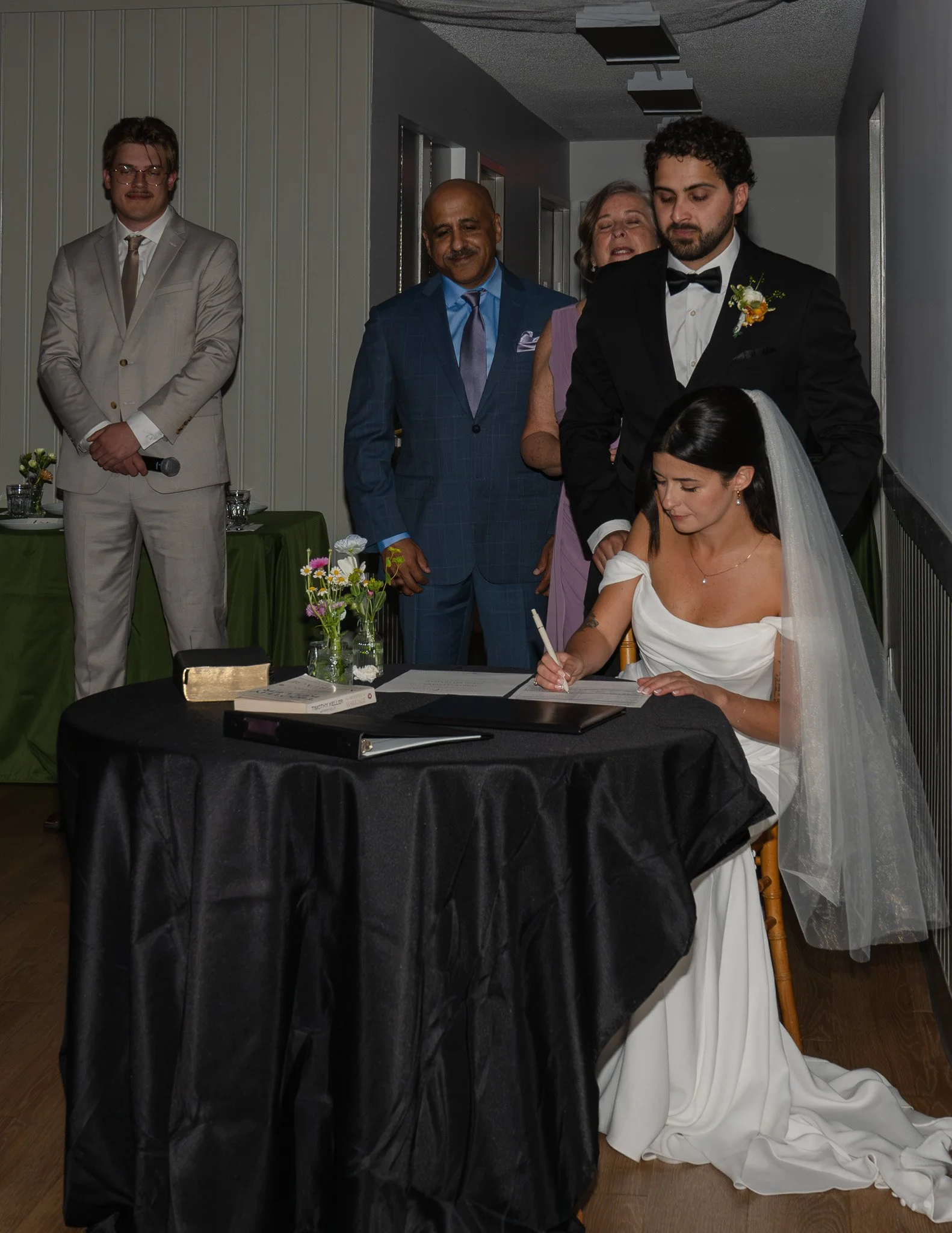 Wedding ceremony with bride signing wedding documents at a table, surrounded by family members and officiant in an indoor venue.
