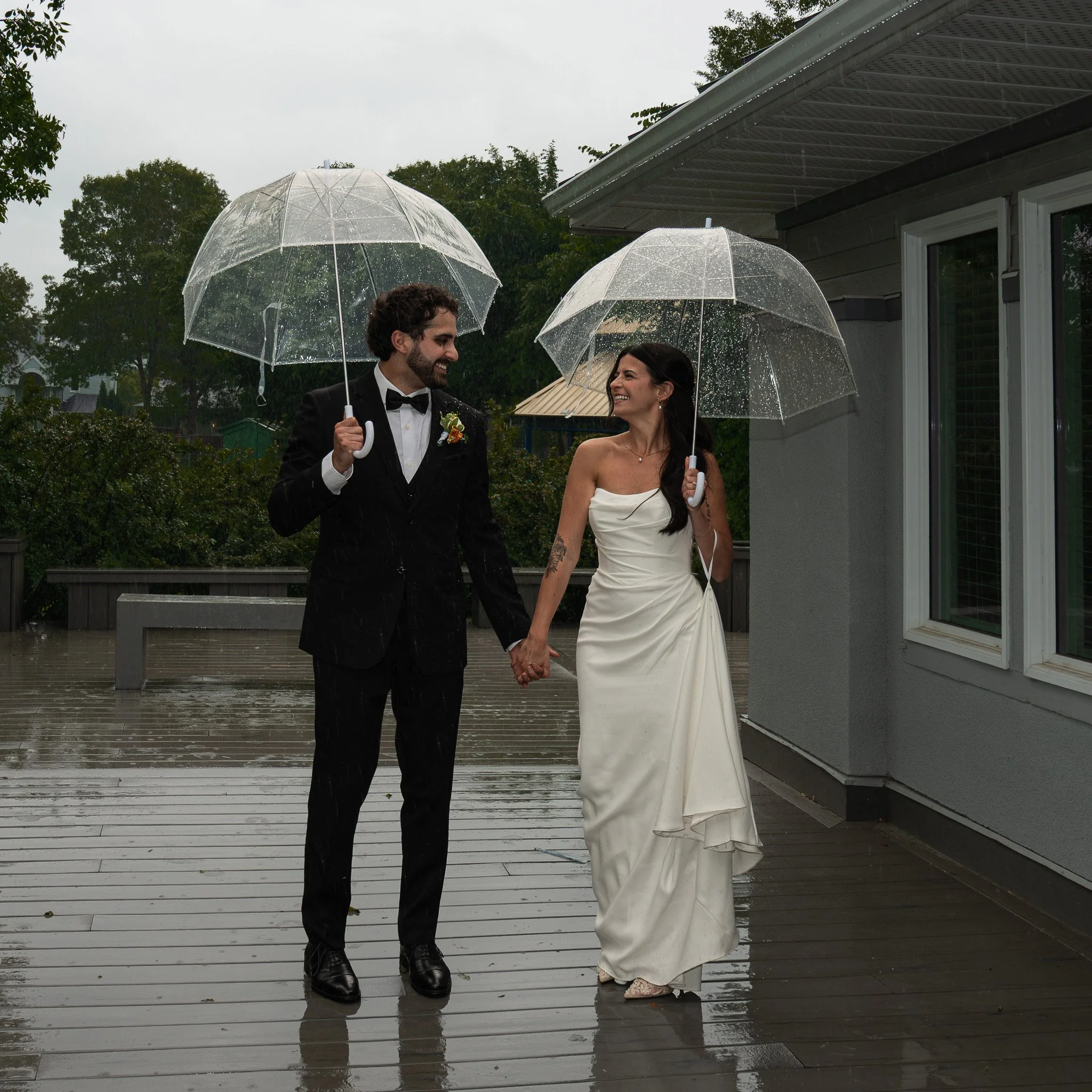 A man and woman dressed in formal wedding attire holding transparent umbrellas in the rain, walking on a wooden deck outside a house with greenery and trees in the background.