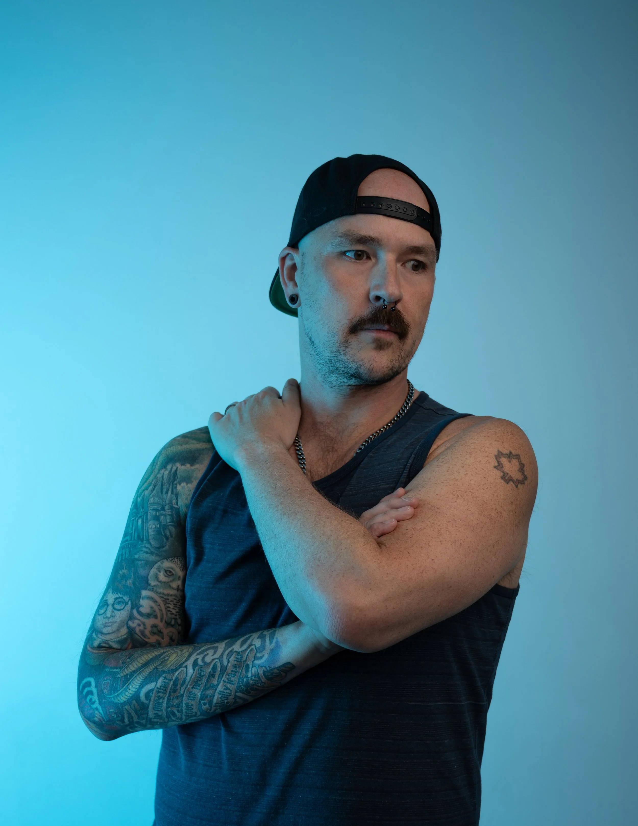 A man with a beard and tattoos on his left arm, wearing a black backwards baseball cap and a black sleeveless shirt, is standing against a gradient blue background, looking down with a serious expression.