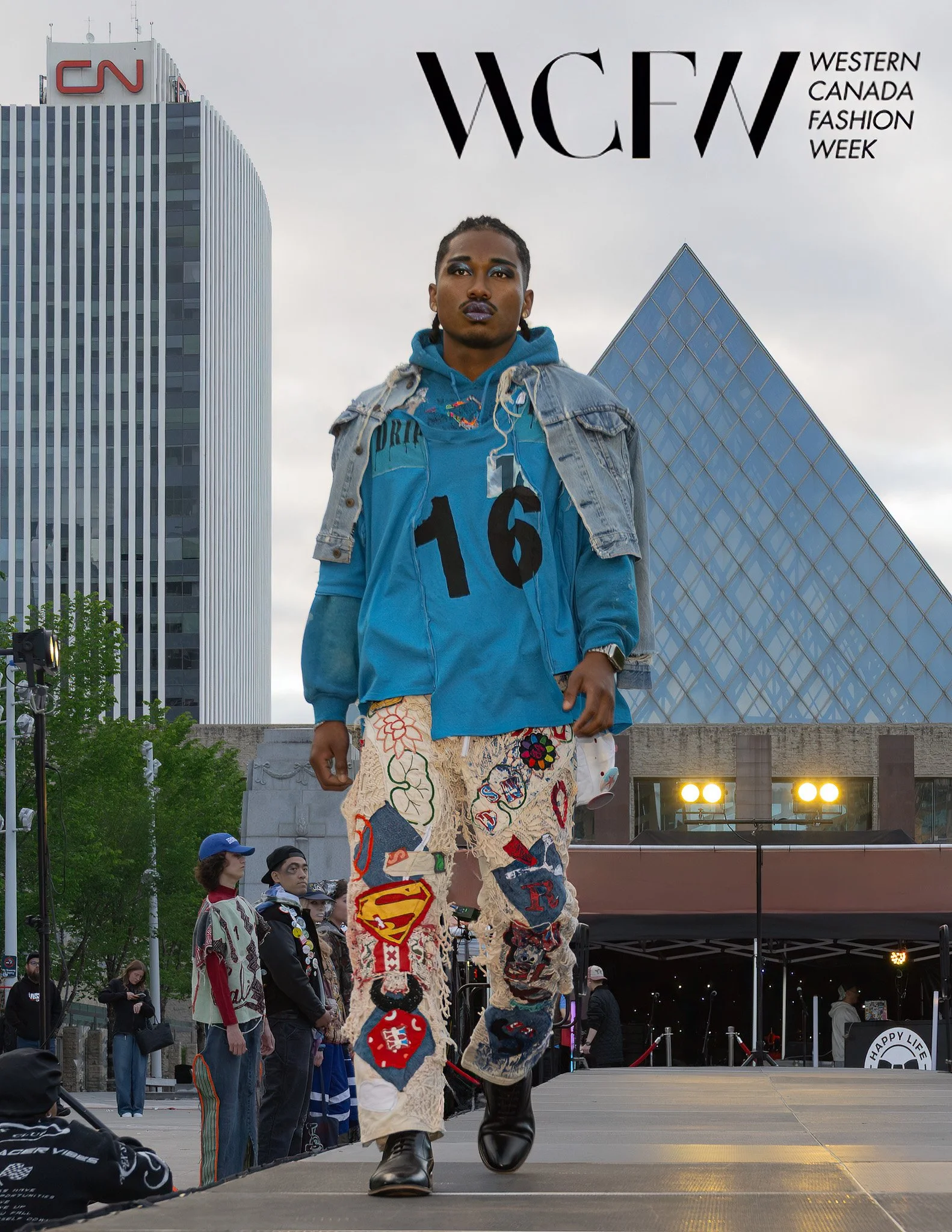 A person on a small stage at Western Canada Fashion Week wearing patchwork pants with college patches, a blue hoodie with the number 16, a denim jacket, and large boots. There are audience members watching and city buildings in the background.