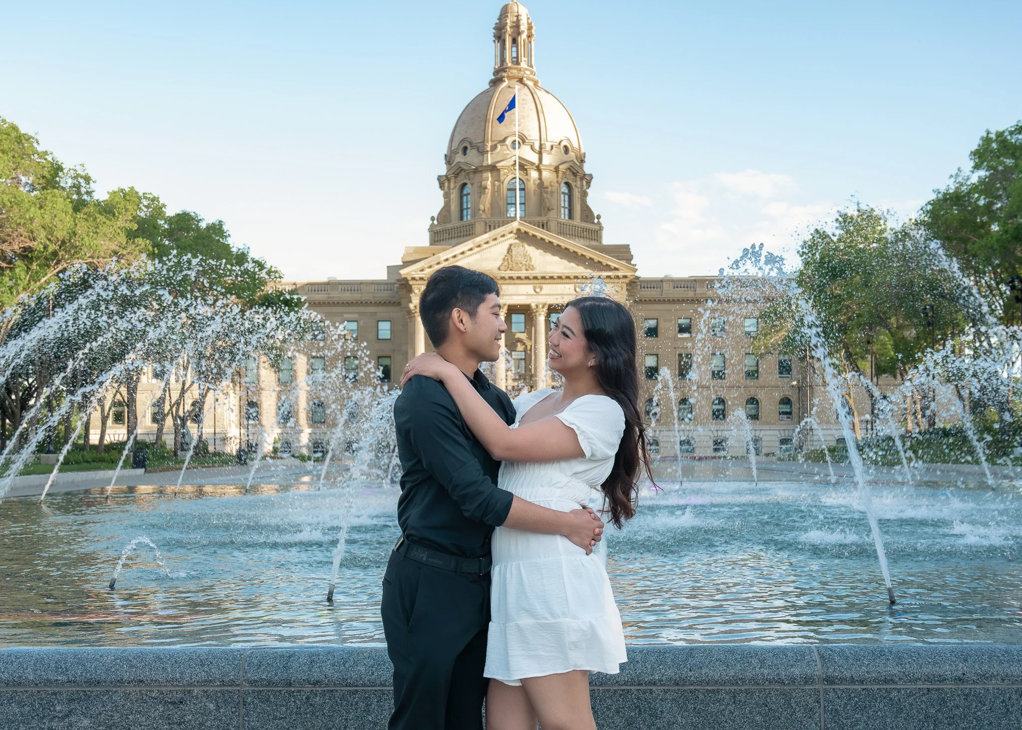 A couple embracing in front of a fountain with water jets, with a historic government building with a dome in the background.