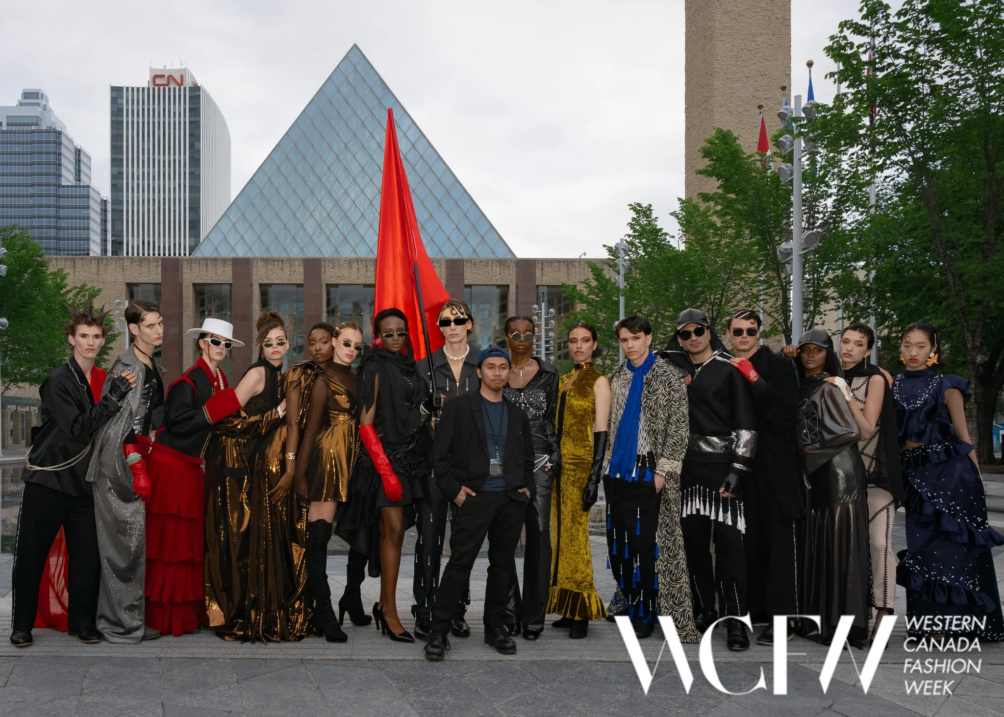Group of diverse fashion models standing together outdoors during daytime, with tall buildings and trees in the background, at Western Canada Fashion Week.