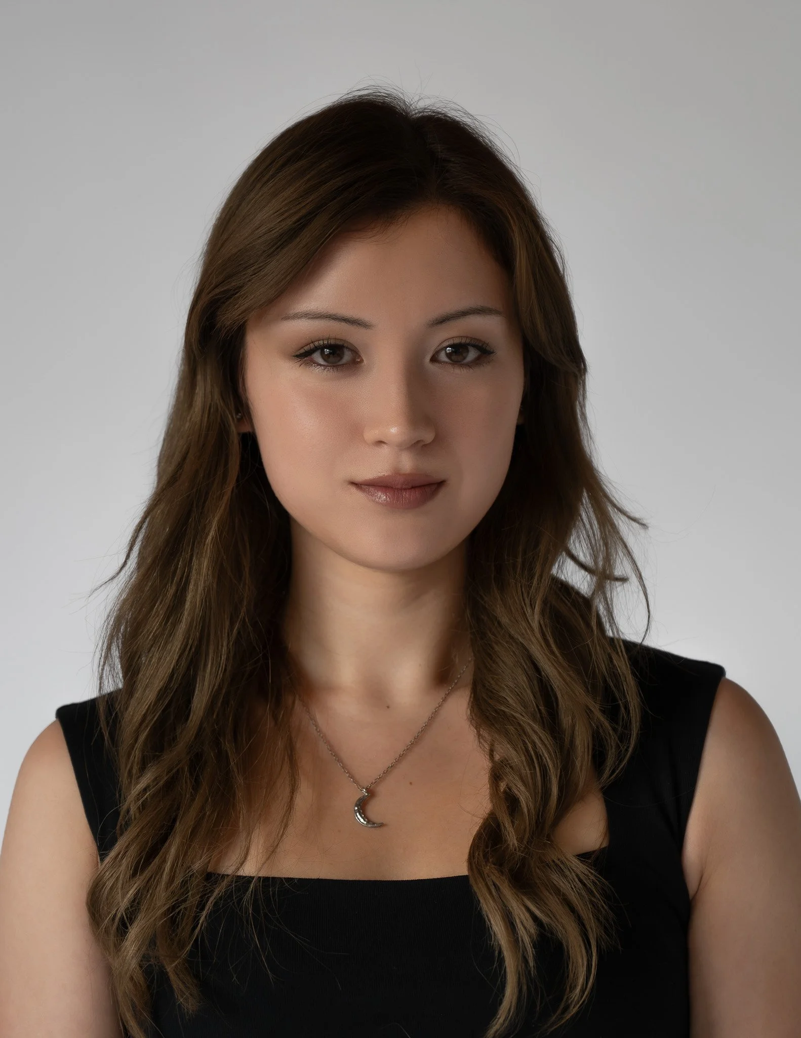 A woman with long wavy brown hair wearing a black sleeveless top, a crescent moon necklace, and a delicate chain.