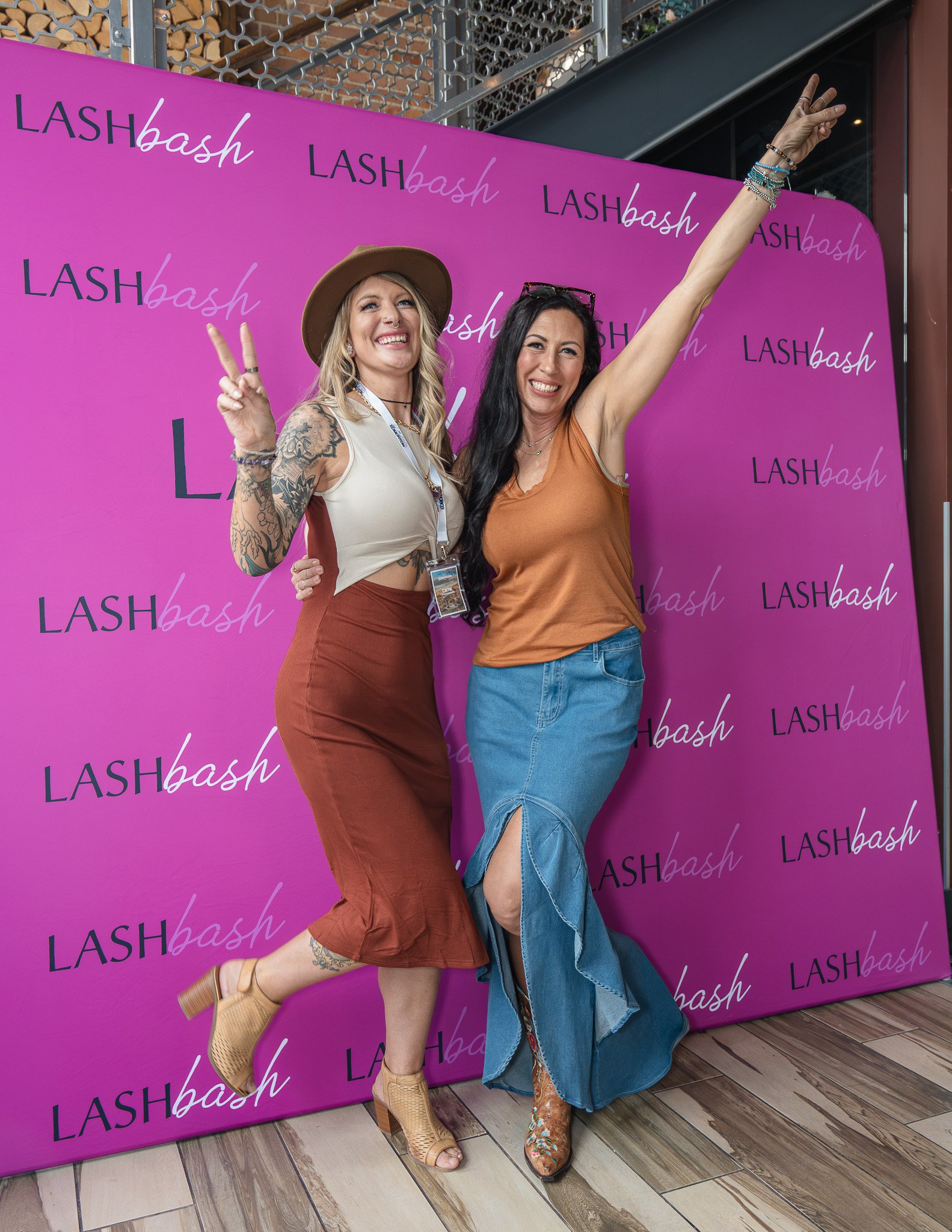 Two women posing happily at a Lash Bash event in front of a purple backdrop with a repeating 'LASH bash' logo pattern. The woman on the left is making a peace sign with her right hand and is wearing a beige hat, sleeveless top, rust-colored skirt, an
