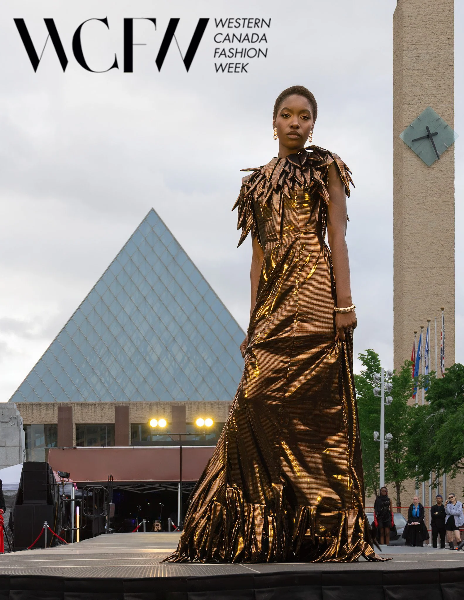 Fashion model in a metallic, gold and black striped dress with a feathered collar walking on runway at Western Canada Fashion Week, with audience and city buildings in background.