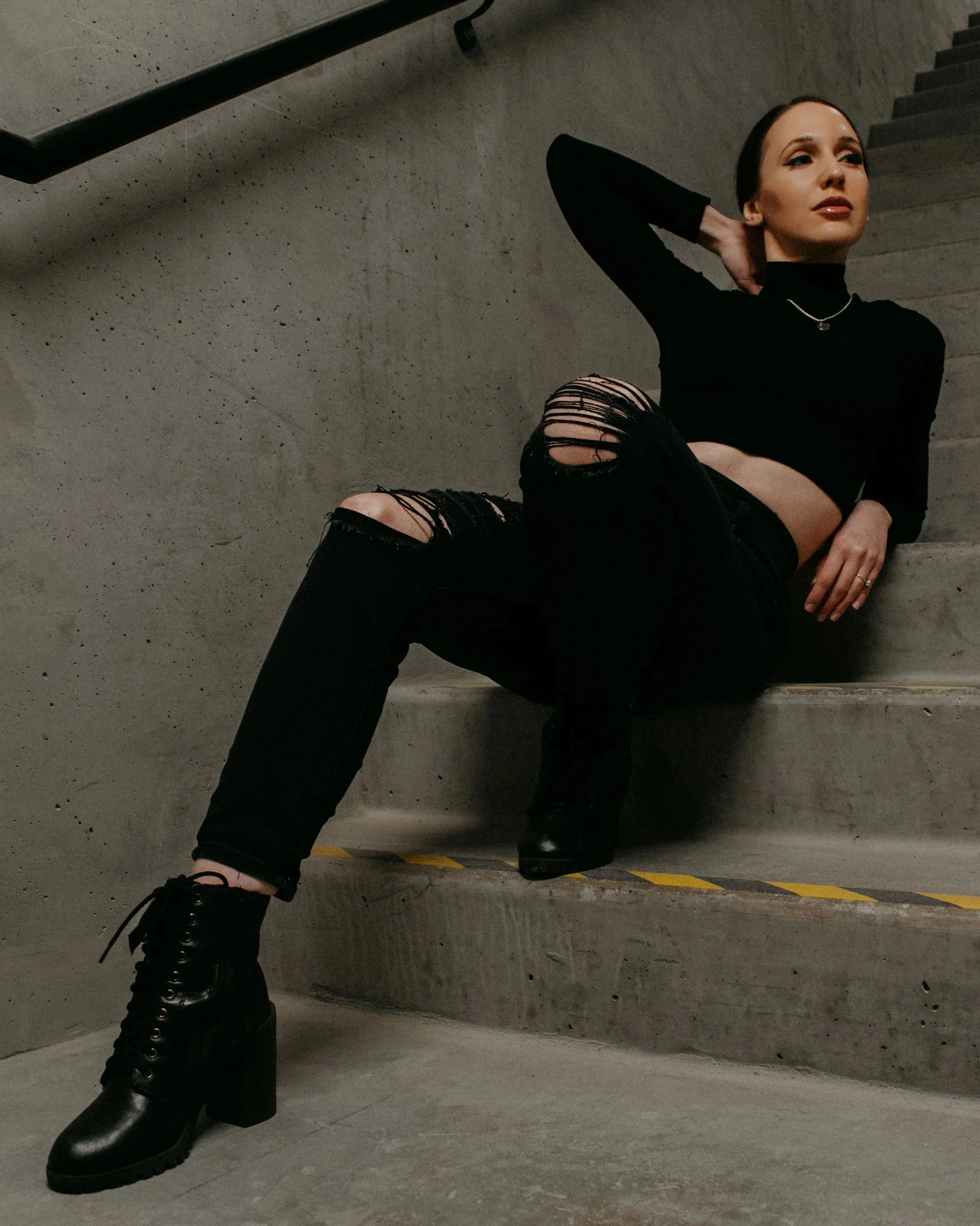 A young woman with dark hair, wearing a black long-sleeve crop top, ripped black jeans, and black heeled lace-up boots, sitting on concrete stairs in an industrial setting with a concrete wall backdrop.