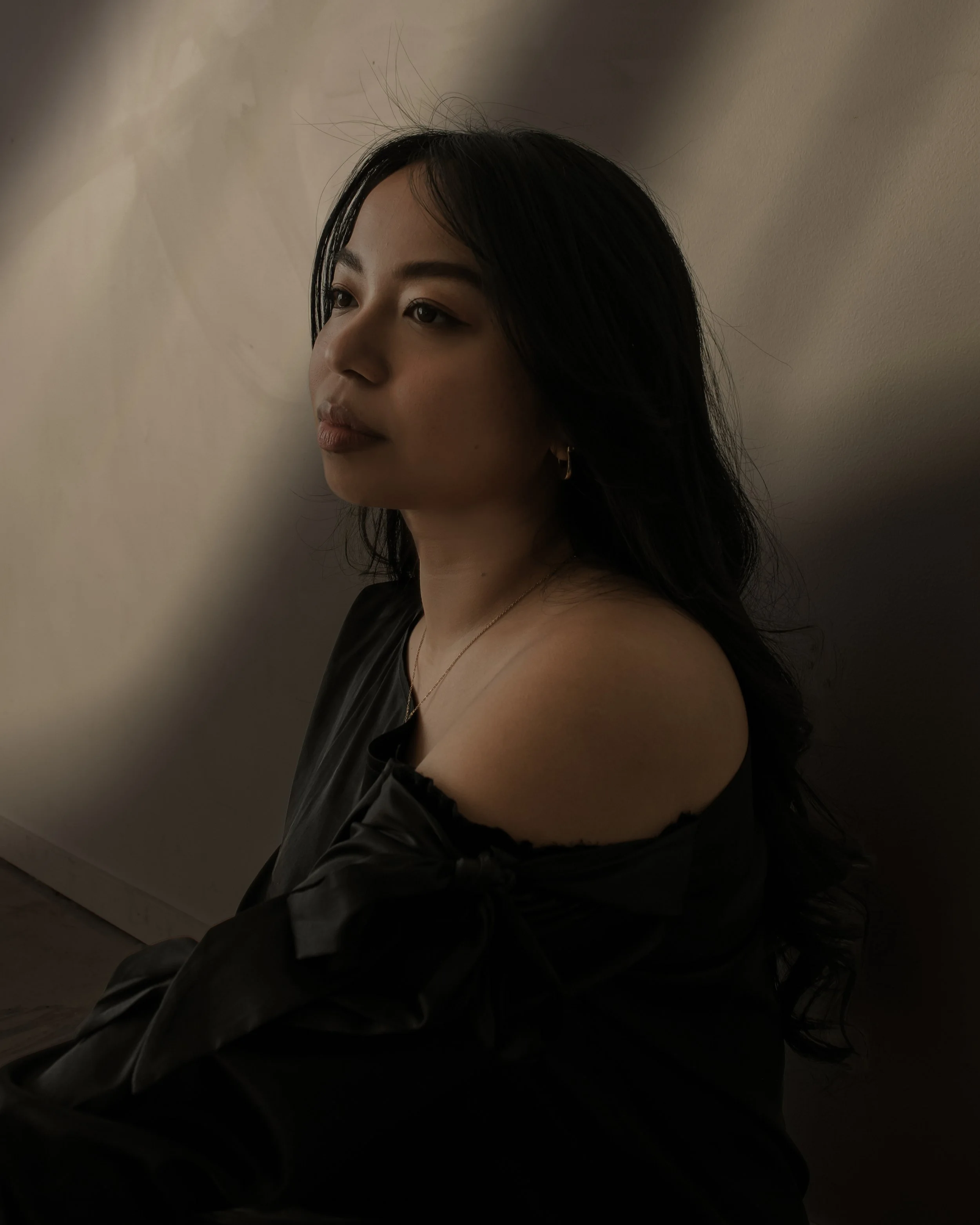 A young woman with long black hair and earrings, wearing an off-shoulder black top, sits against a beige wall with soft lighting.