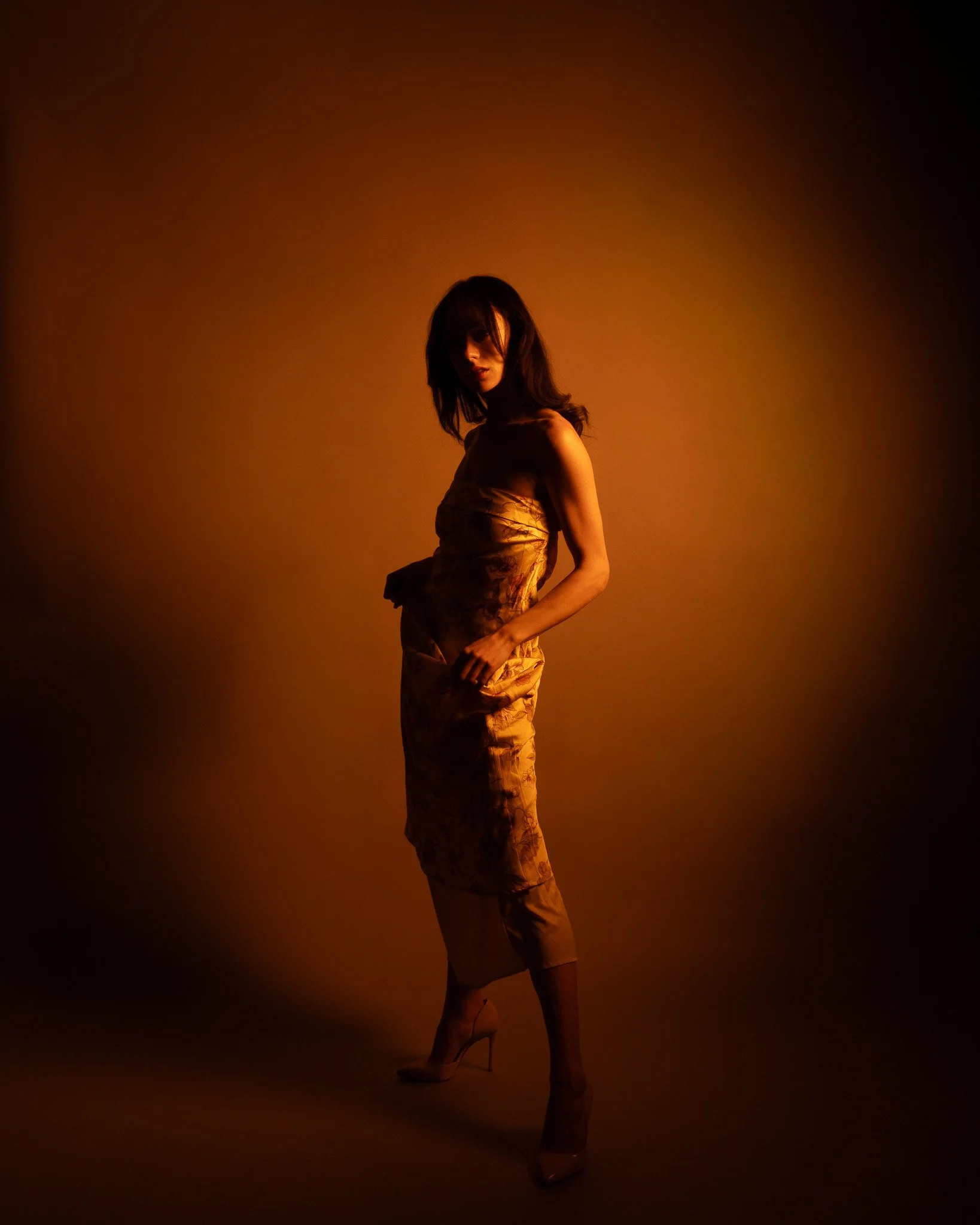 A woman with dark hair wearing a beige dress and high heels stands against a warm gradient background, with dramatic lighting casting shadows on her face and body.
