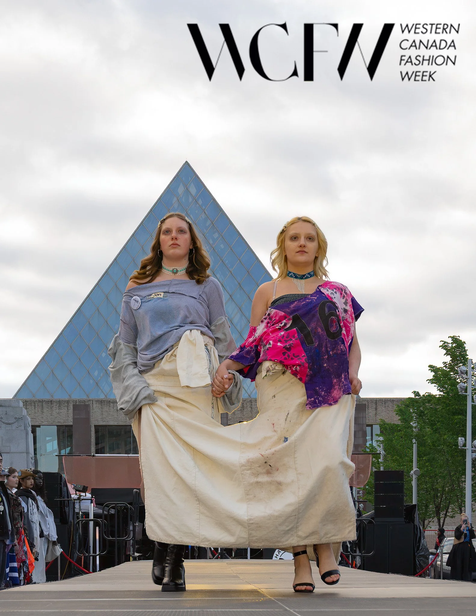 Two women walking on a runway during Western Canada Fashion Week; one in boots and the other in sandals, with a pyramid-shaped building and cloudy sky in the background