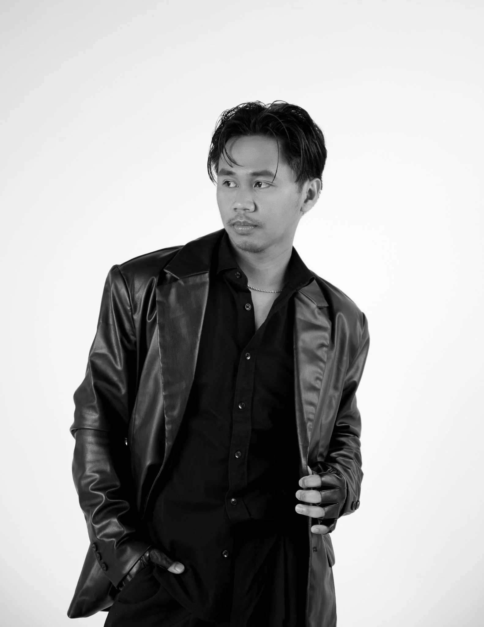 A young man with dark hair styled back, wearing a black leather jacket over a black shirt, gazing to his left while standing against a plain white background.