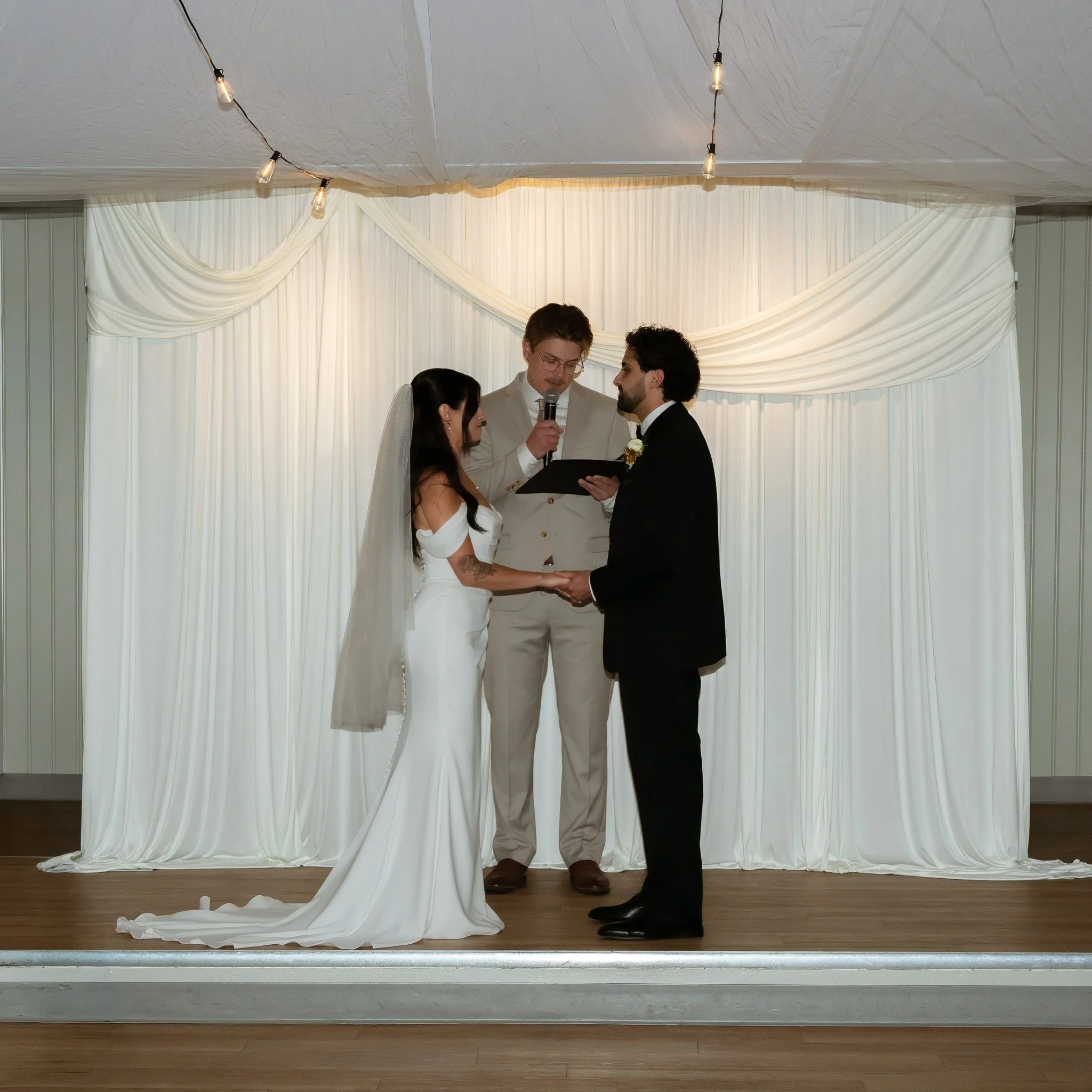 A wedding ceremony taking place on a stage with a white backdrop and draped fabric. The officiant is reading from a book while holding a microphone. The couple is standing before him, holding hands, with the bride in a white wedding dress and the gro