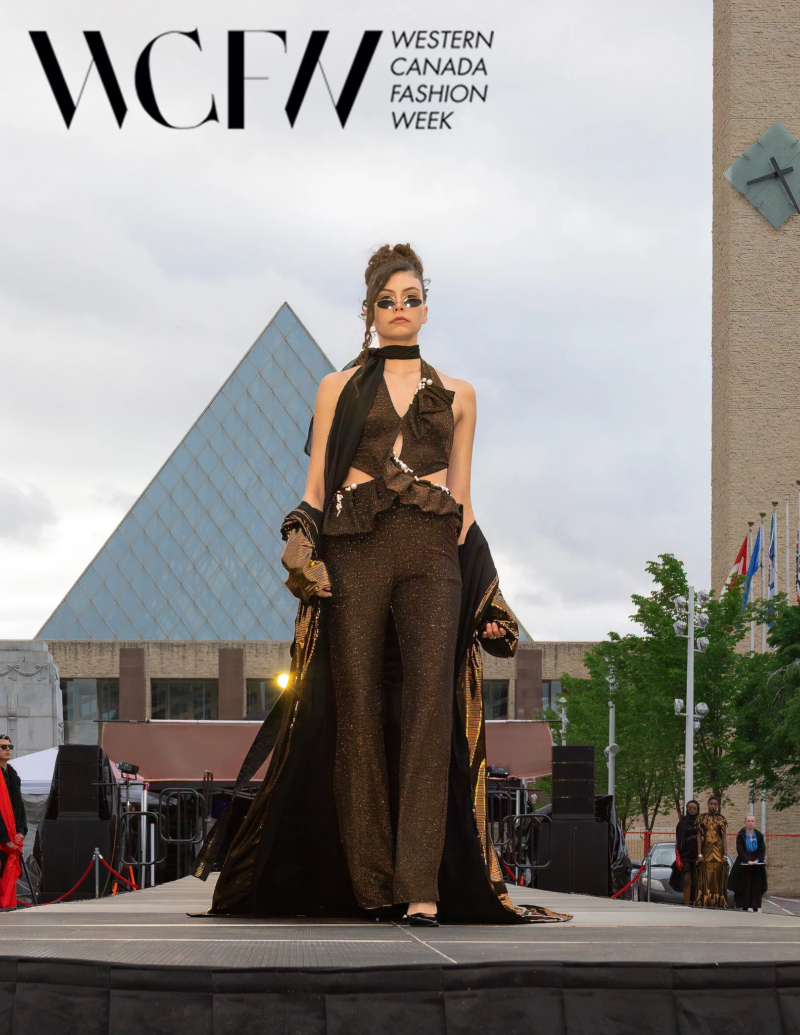 Fashion model walking on stage during Western Canada Fashion Week, wearing a black and gold outfit with sheer pants and a matching jacket, with the event banner and audience in the background.