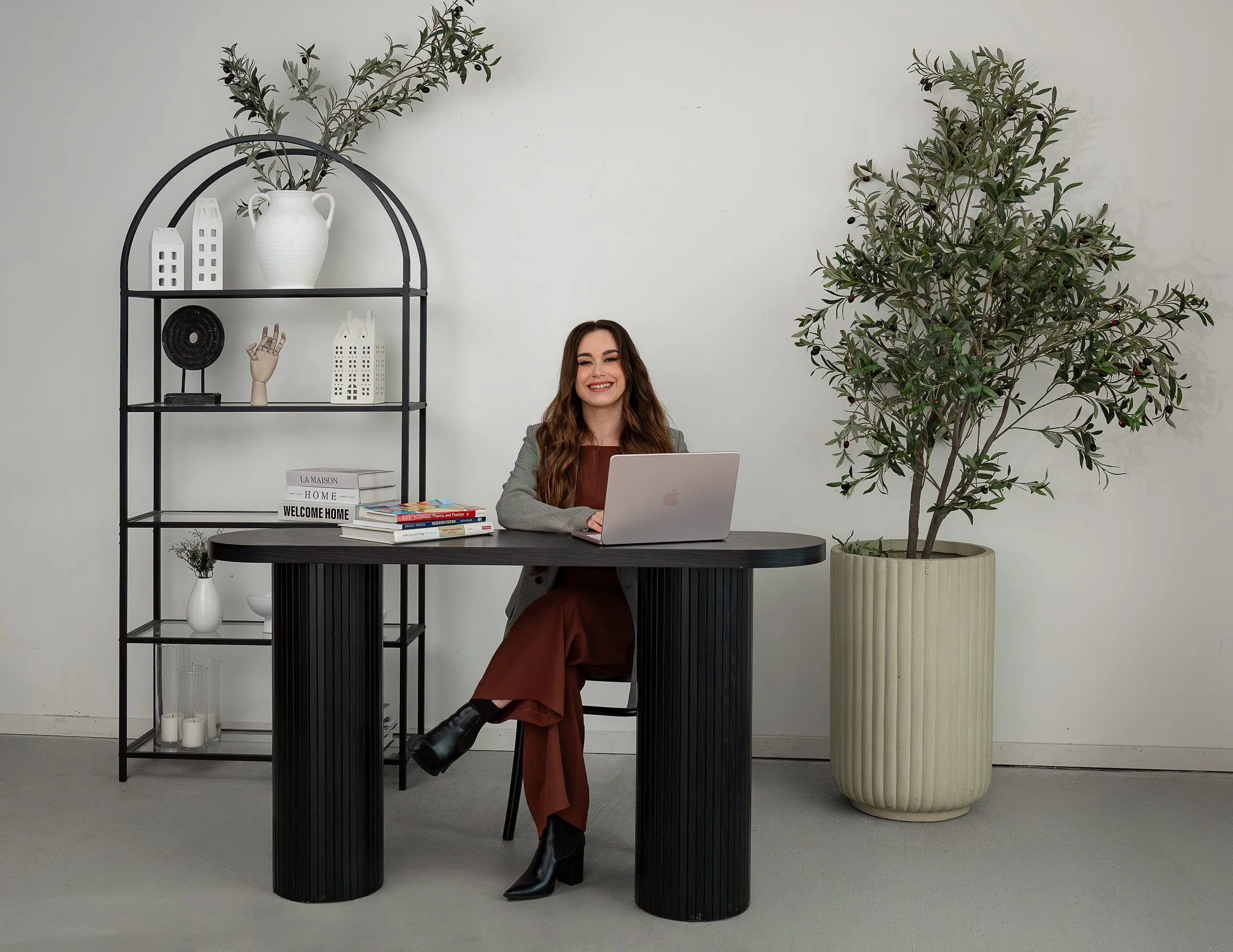 A woman sitting at a black table with a laptop, smiling in an office setting with large plants and decorative shelves.