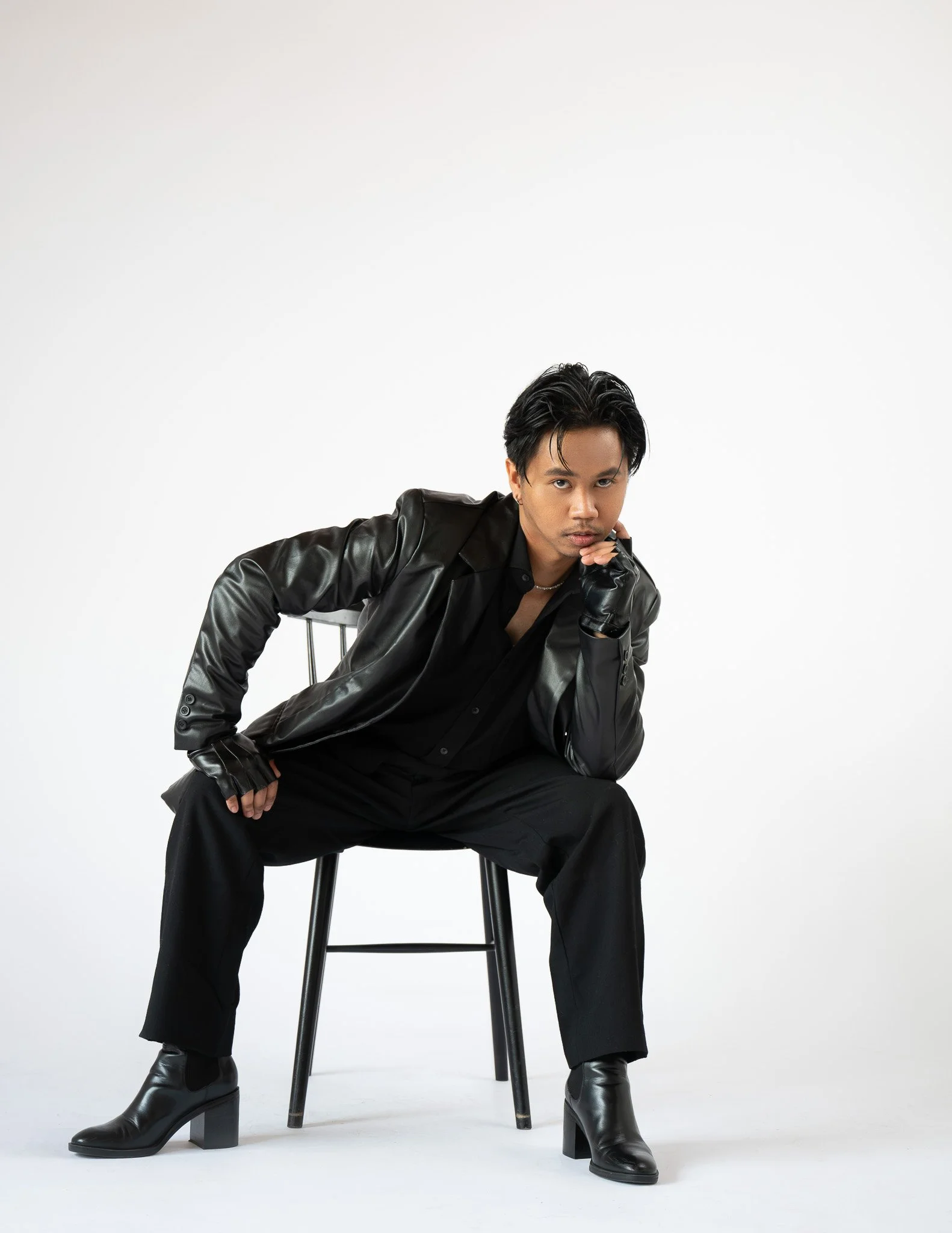 A young man with dark hair, wearing a black leather jacket, black shirt, black gloves, and black high boots, seated on a chair, looking at the camera with a serious expression, against a plain white background.