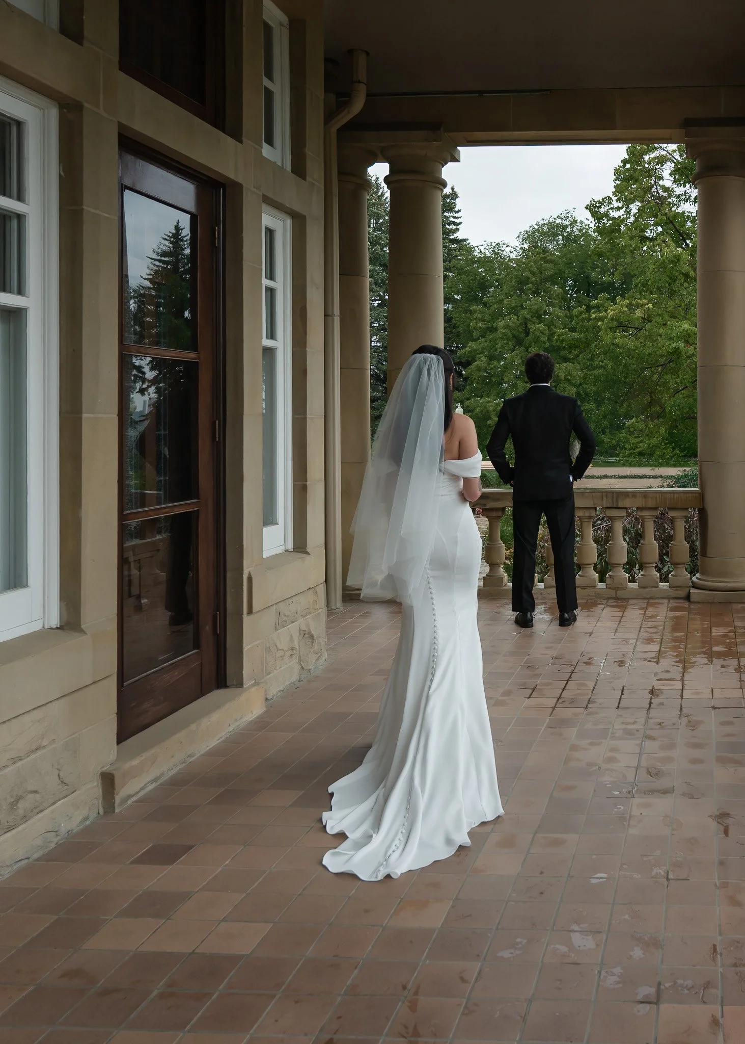 A bride and groom standing on a terrace with stone columns, trees and an overcast sky in the background, waiting for their wedding ceremony.