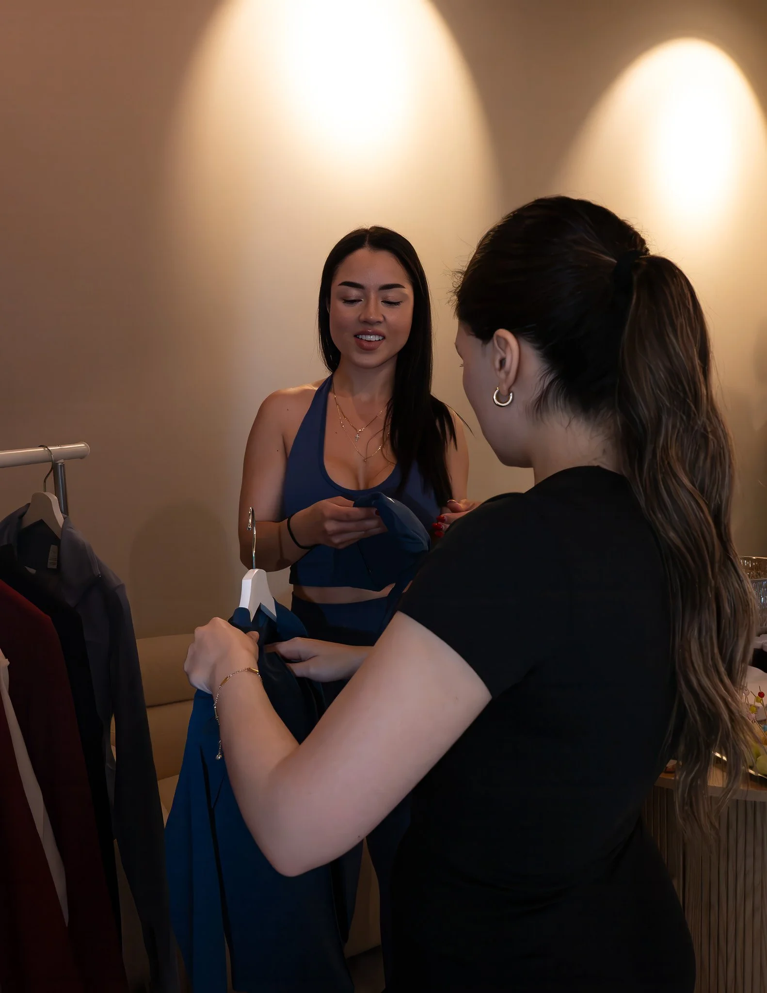 Two women are indoors, one in a blue sports top and the other in a black shirt, engaging with clothing items on a hanger. The woman in blue has long dark hair, and the woman in black has wavy hair tied back. Warm lighting is cast on the wall behind t