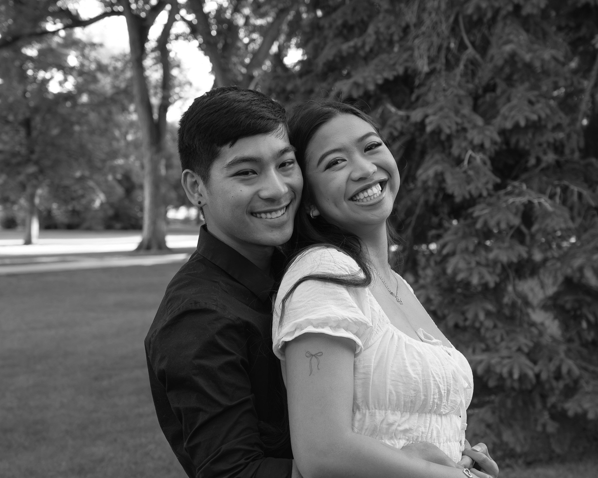 A smiling couple, a man and a woman, standing close together outdoors in a park with trees in the background. The photograph is in black and white.