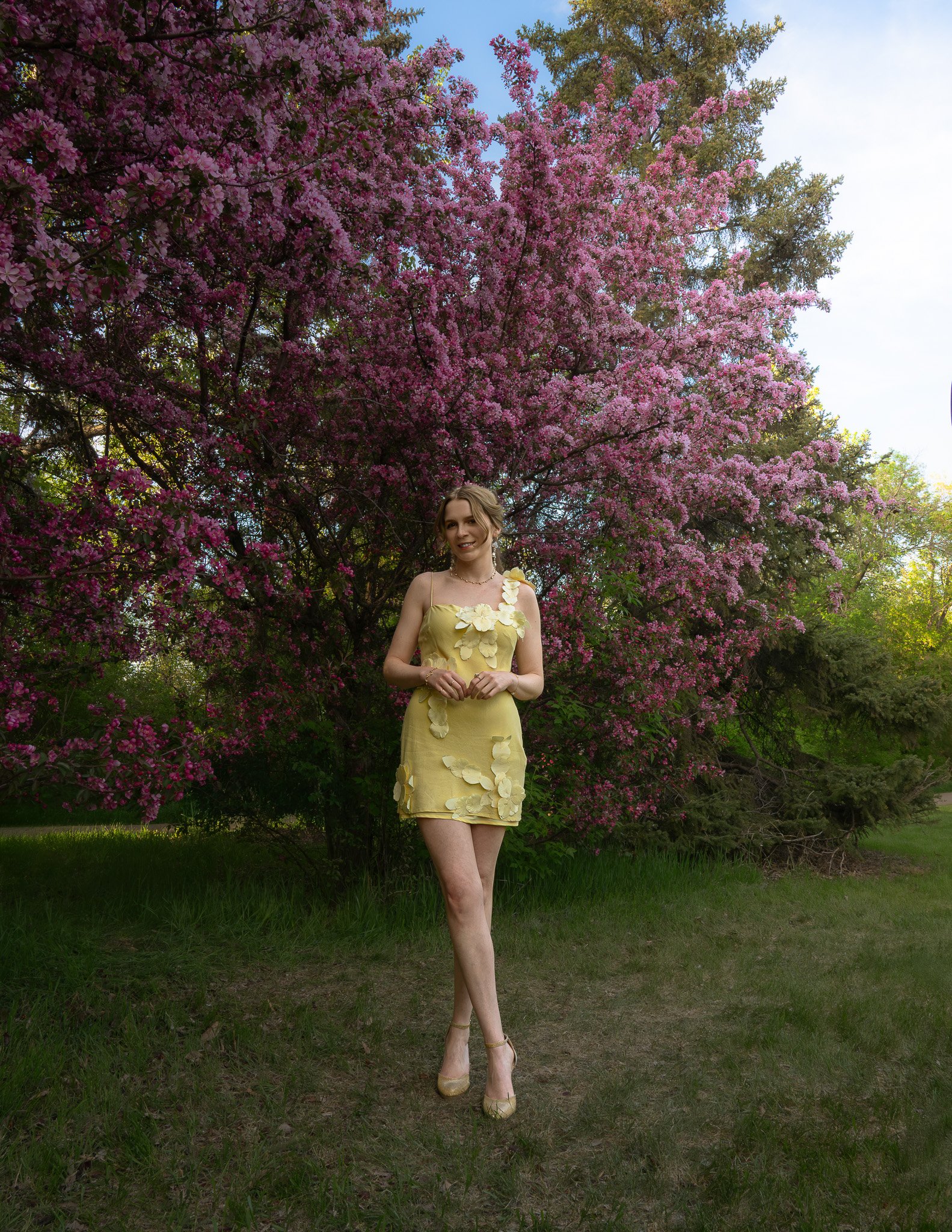 A woman in a yellow dress with floral appliqué details stands outdoors in front of a bush with pink blossoms. She is smiling, with her hair styled in loose waves, wearing heels, and surrounded by lush greenery and trees.