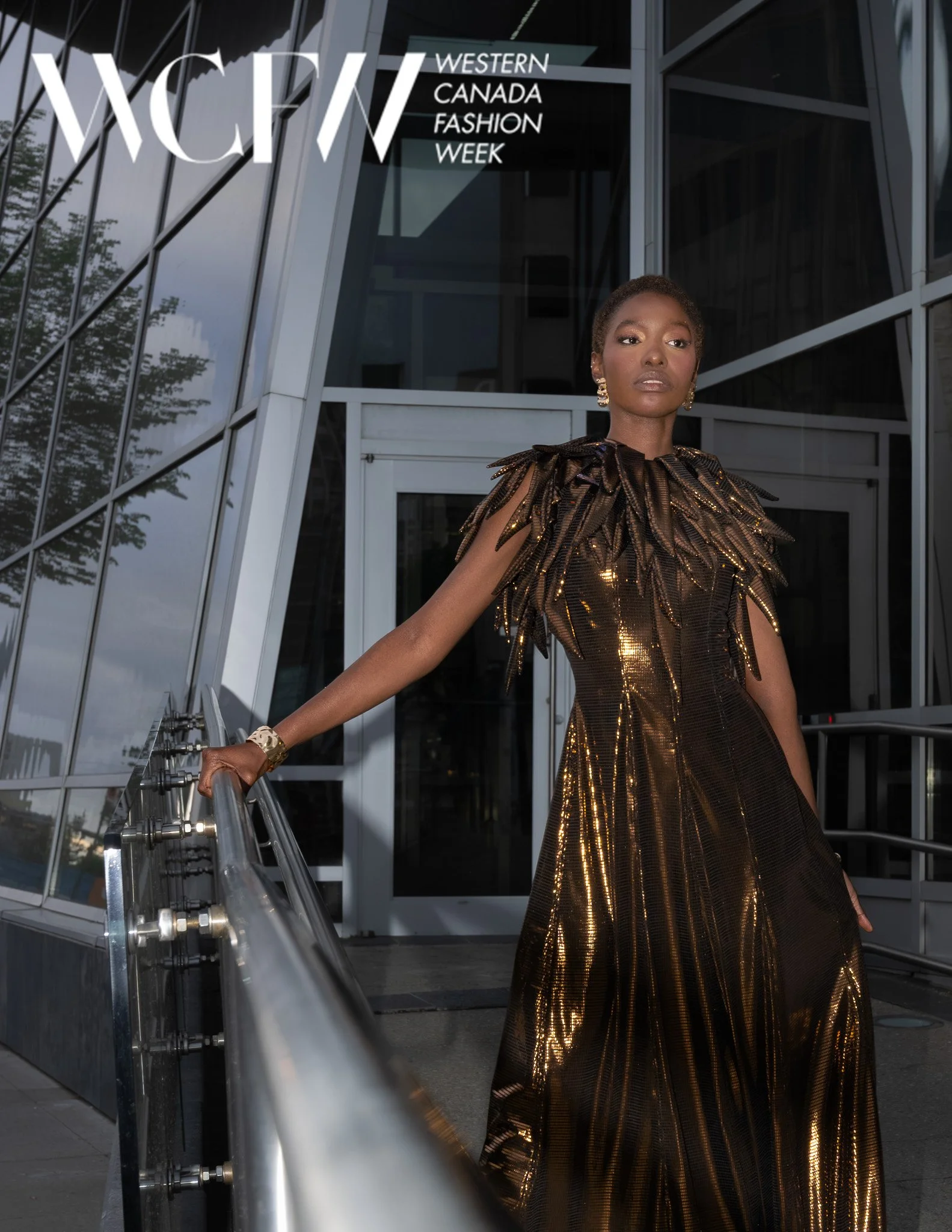 A woman in a shimmering gold, metallic gown with feather-like details stands in front of a modern glass building during Western Canada Fashion Week. She is accessorized with earrings and a bracelet.