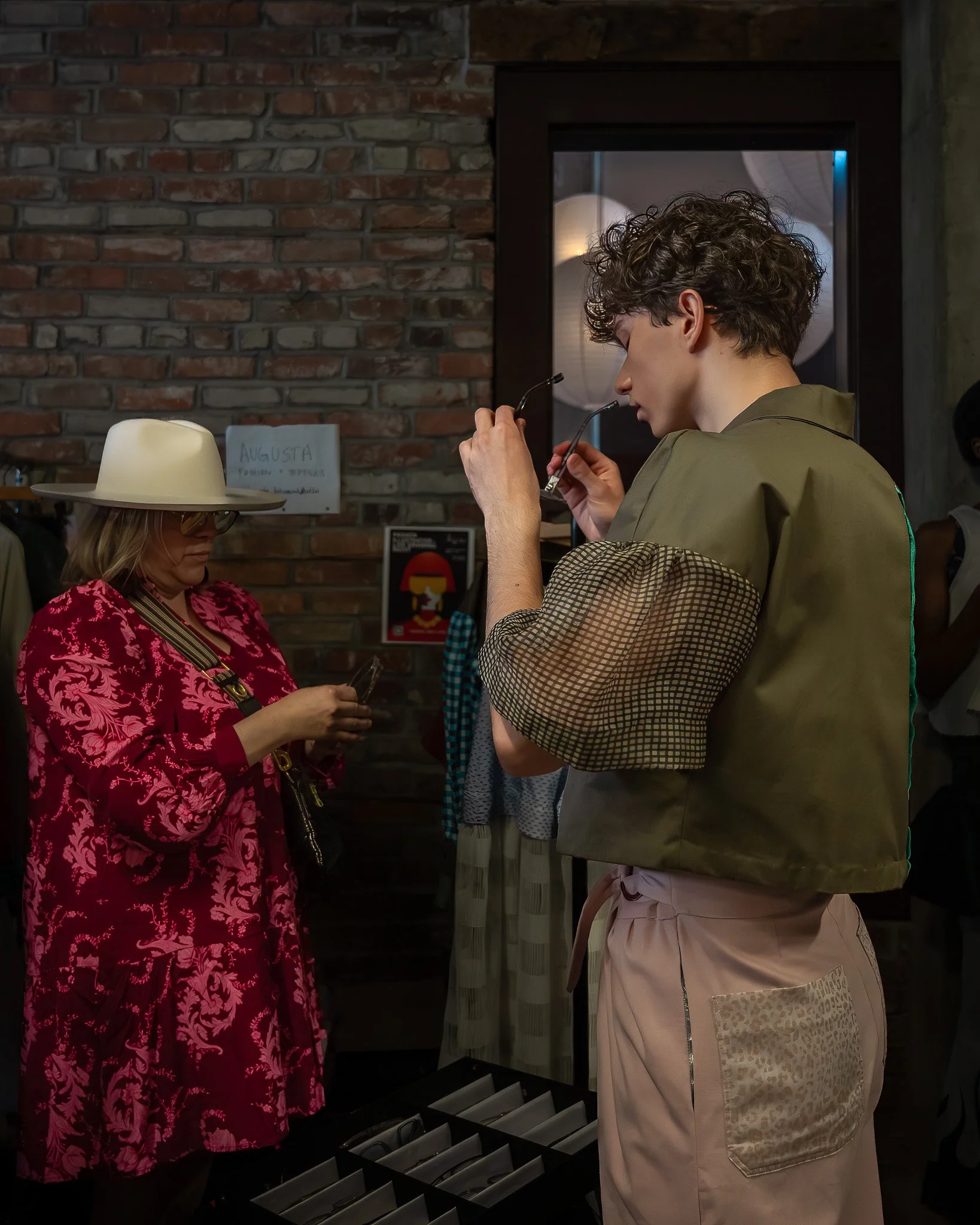 A young man wearing light-colored pants with leopard print patch on pocket and olive green jacket is trying on eyeglasses in a store. A woman with a wide-brimmed hat and red floral dress stands nearby. The background features a brick wall with a sign