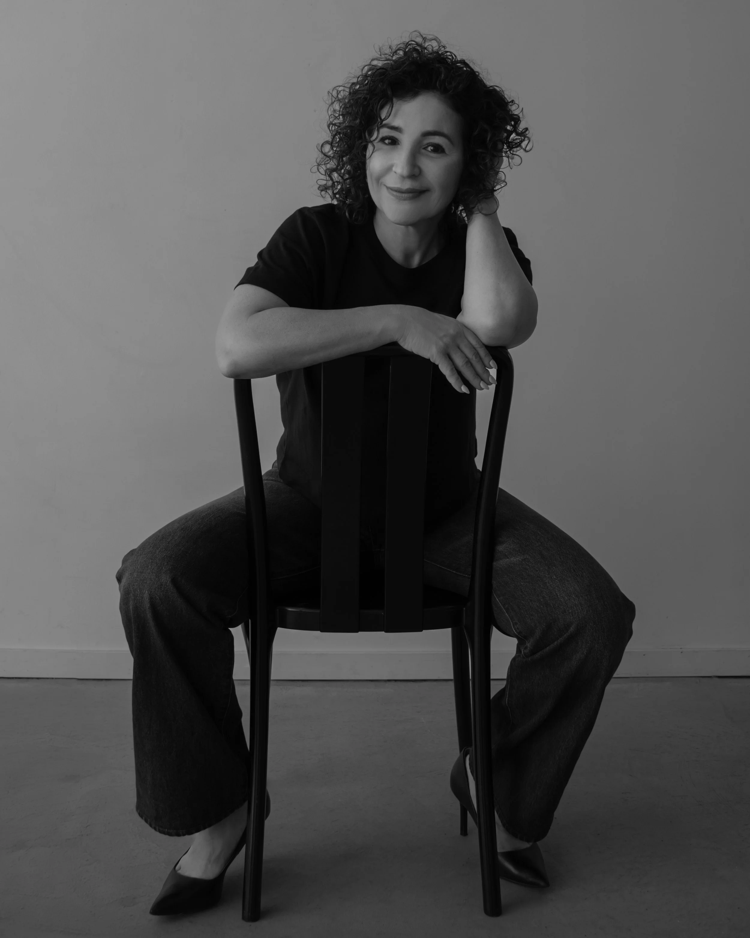 A woman with curly hair sitting on a chair with her legs spread, wearing a black t-shirt and jeans, smiling at the camera in a black and white photo.