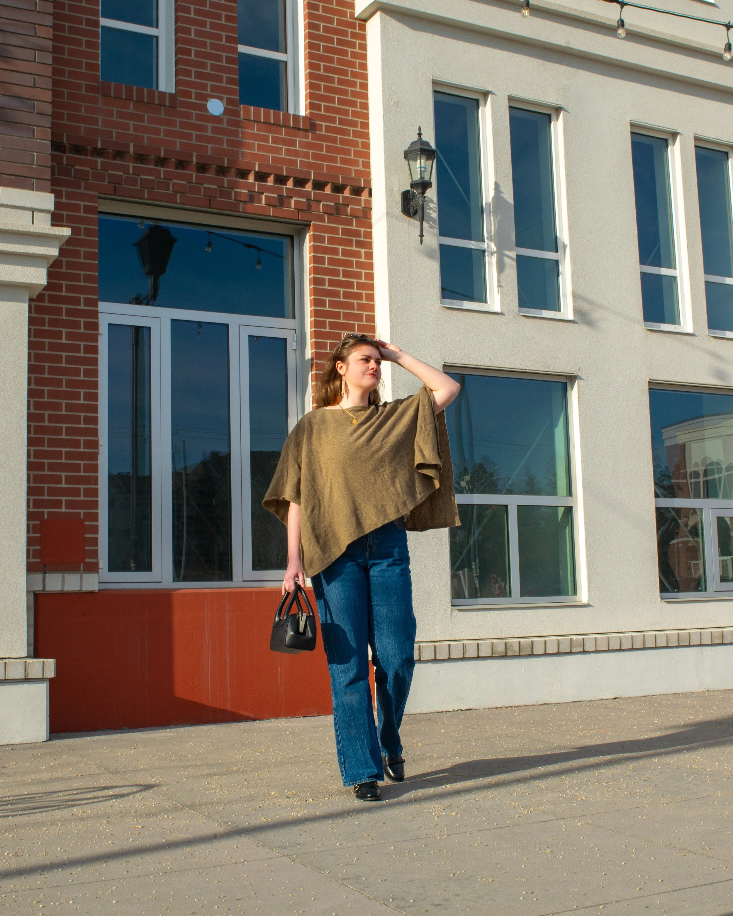 A woman wearing a brown poncho, blue jeans, and black shoes standing outside in front of a modern building with large windows and brick and white walls. She is holding a black handbag and shielding her eyes from the sun with her hand, looking into the distance. There are string lights above her and shadows on the ground.