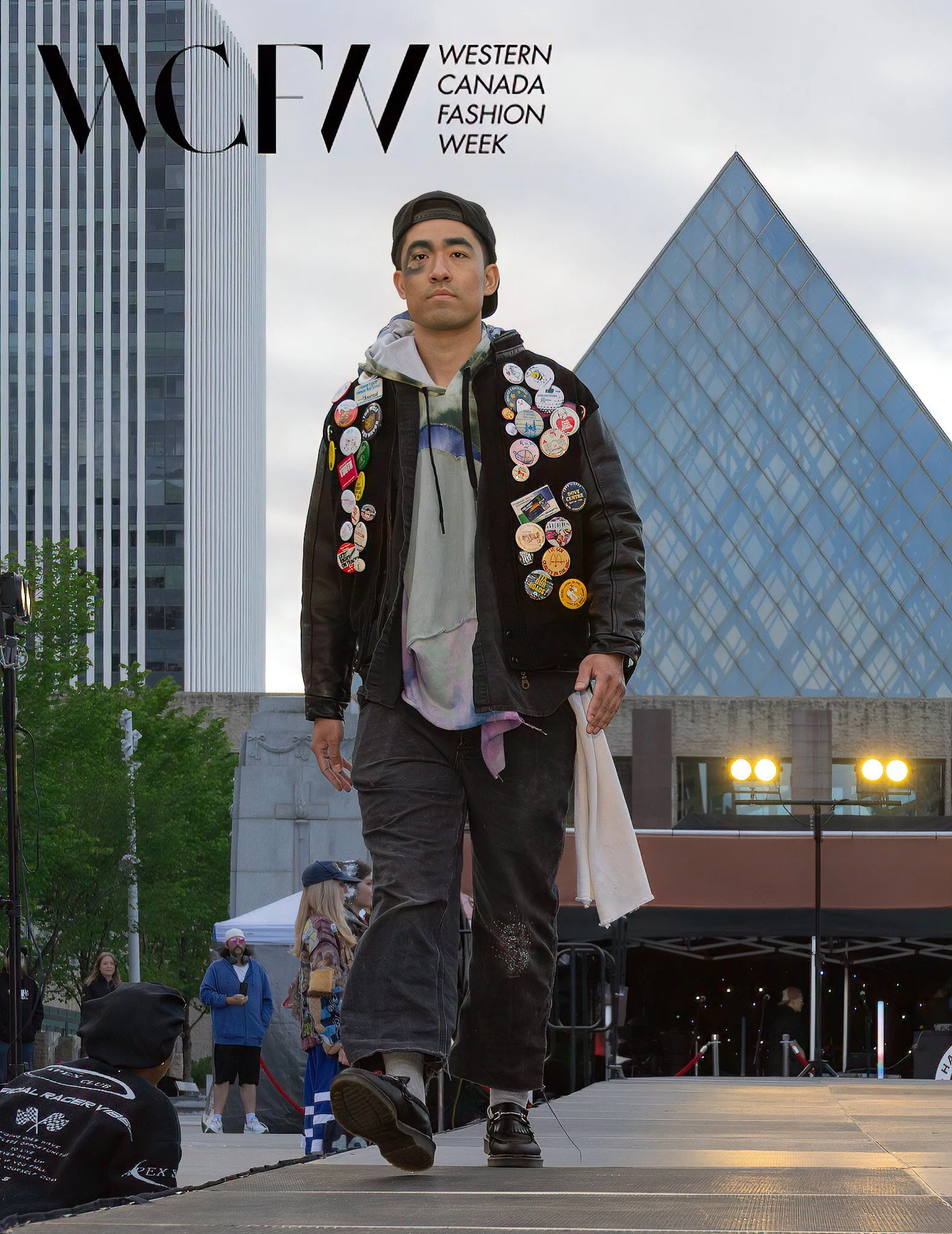 Street scene with a man walking on a stage at Western Canada Fashion Week, wearing a jacket covered in numerous buttons and pins. Background includes modern buildings, and several onlookers watching the event.
